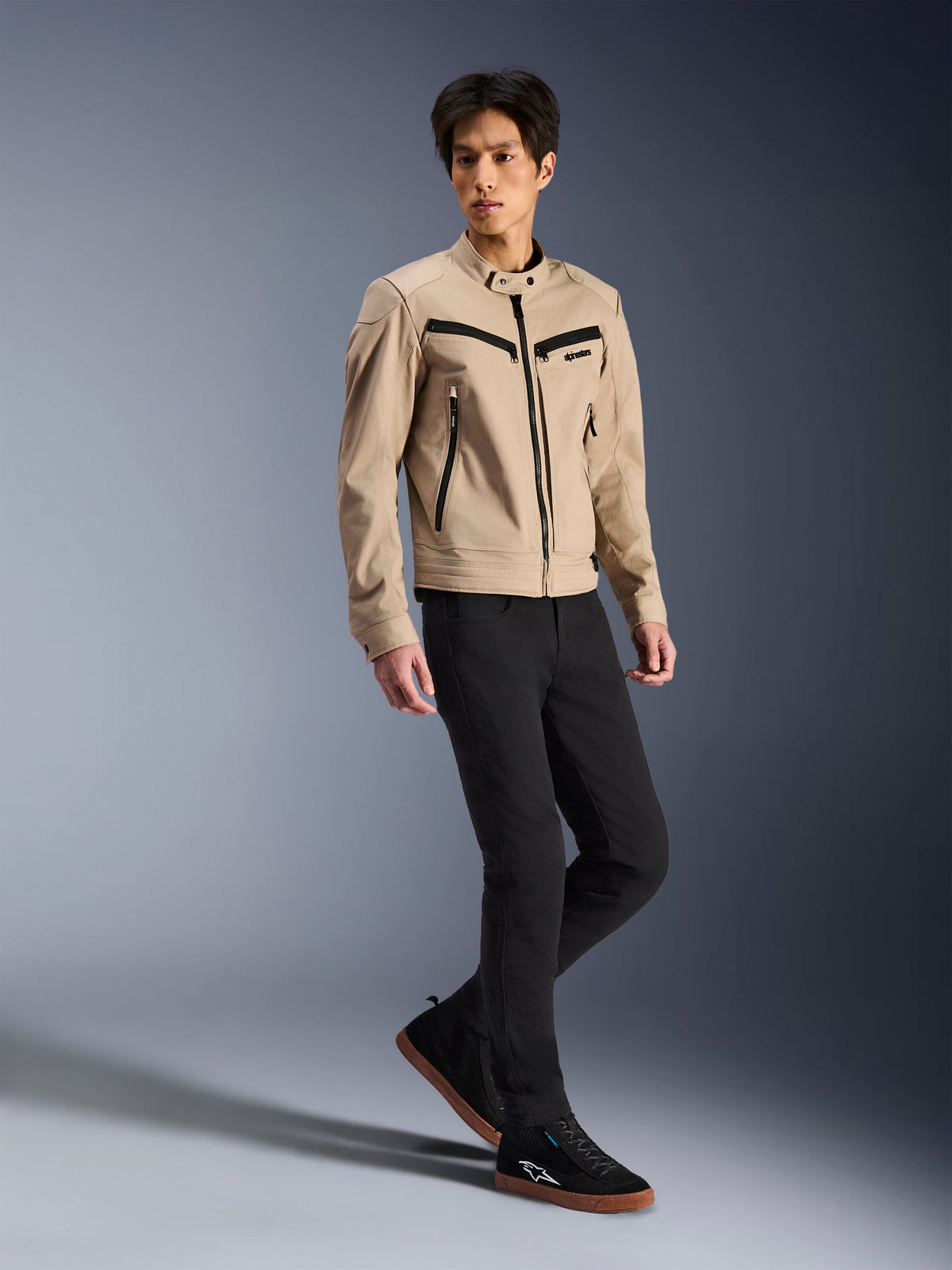 Alpinestars Bruiser Jacket, miejska kurtka motocyklowa, piaskowa, prezentowana przez modela w czarnych spodniach typu slim i czarnych trampkach, tekstylna konstrukcja inspirowana odzieżą roboczą z czarnymi akcentami suwaków, współczesny miejski styl uliczny.