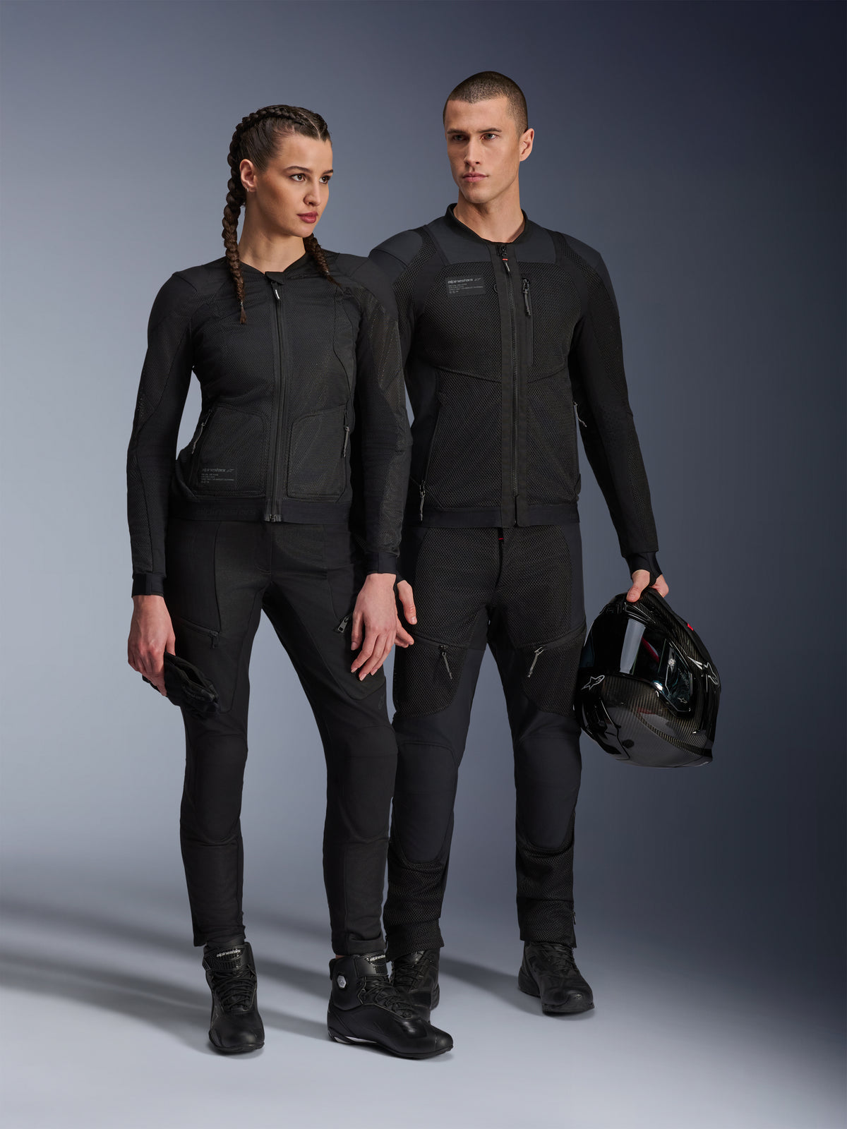 Kurtka Alpinestars Stella Troop-Air, mężczyzna i kobieta w pasującej do siebie, całkowicie czarnej technicznej odzieży motocyklowej z siatkową konstrukcją zapewniającą przepływ powietrza, czarne spodnie i buty, mężczyzna trzymający czarny kask, miejska odzież motocyklowa