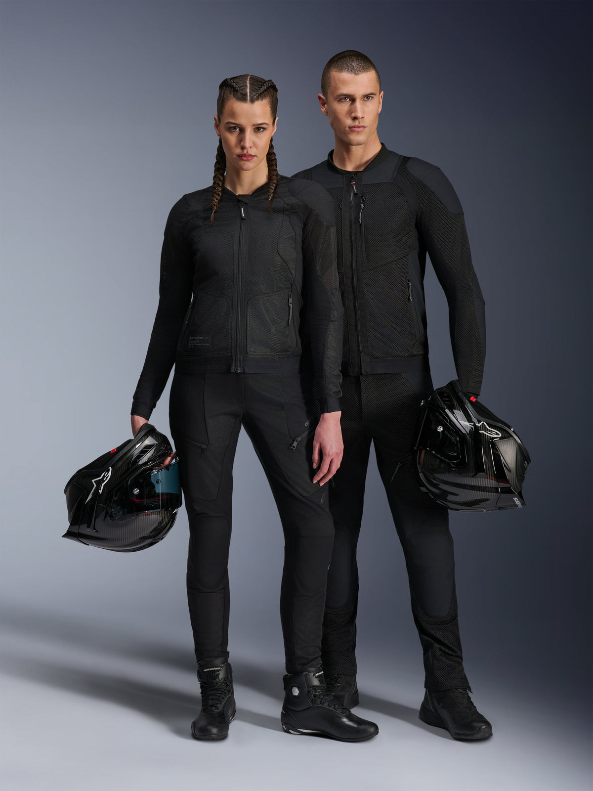 Modele, mężczyzna i kobieta, ubrani w czarne kurtki Alpinestars Troop-Air Pro, techniczną siatkową odzież motocyklową z protektorami w zestawieniu z pasującymi czarnymi spodniami i butami, każdy trzymający czarny kask, prezentujący skoordynowaną miejską odzież motocyklową.