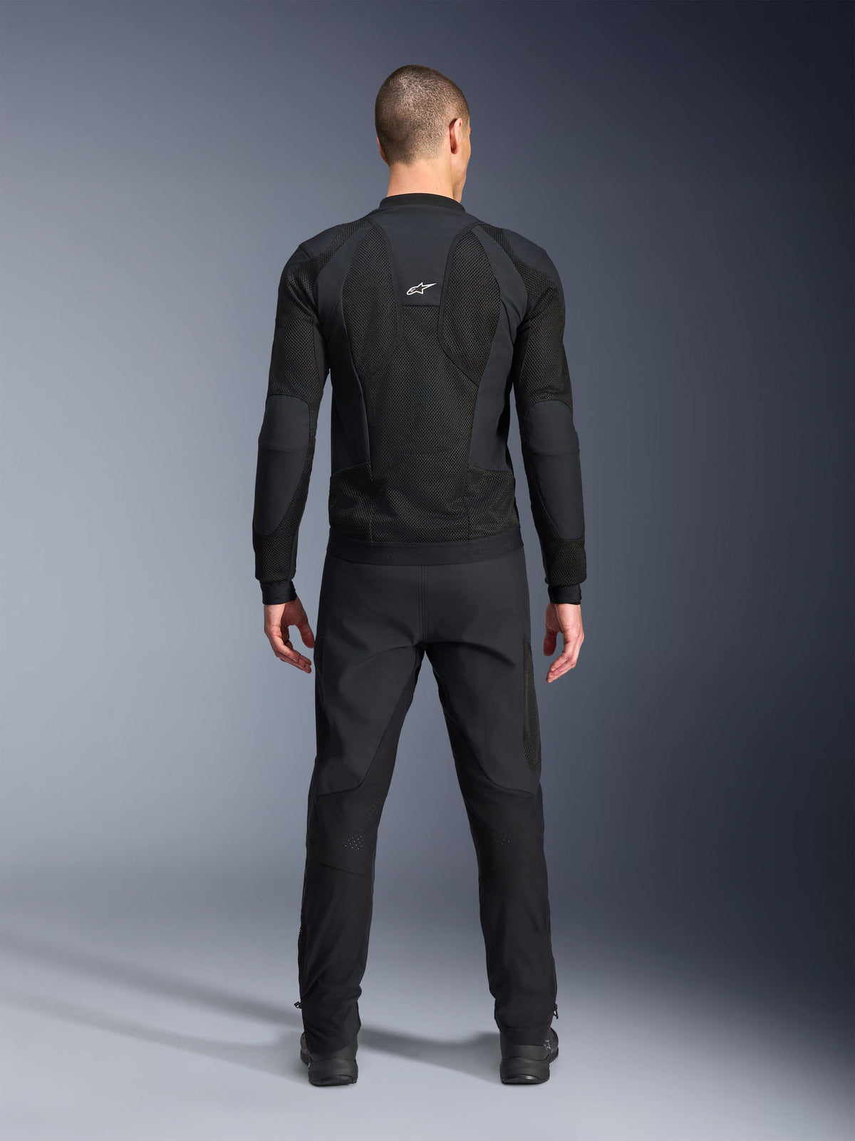 Alpinestars Troop-Air Pro Jacket, widok z tyłu, czarna, techniczna motocyklowa kurtka z dużymi panelami wentylacyjnymi z siatki i białym logo Alpinestars, zestawiona z czarnymi spodniami, nowoczesna miejska odzież motocyklowa