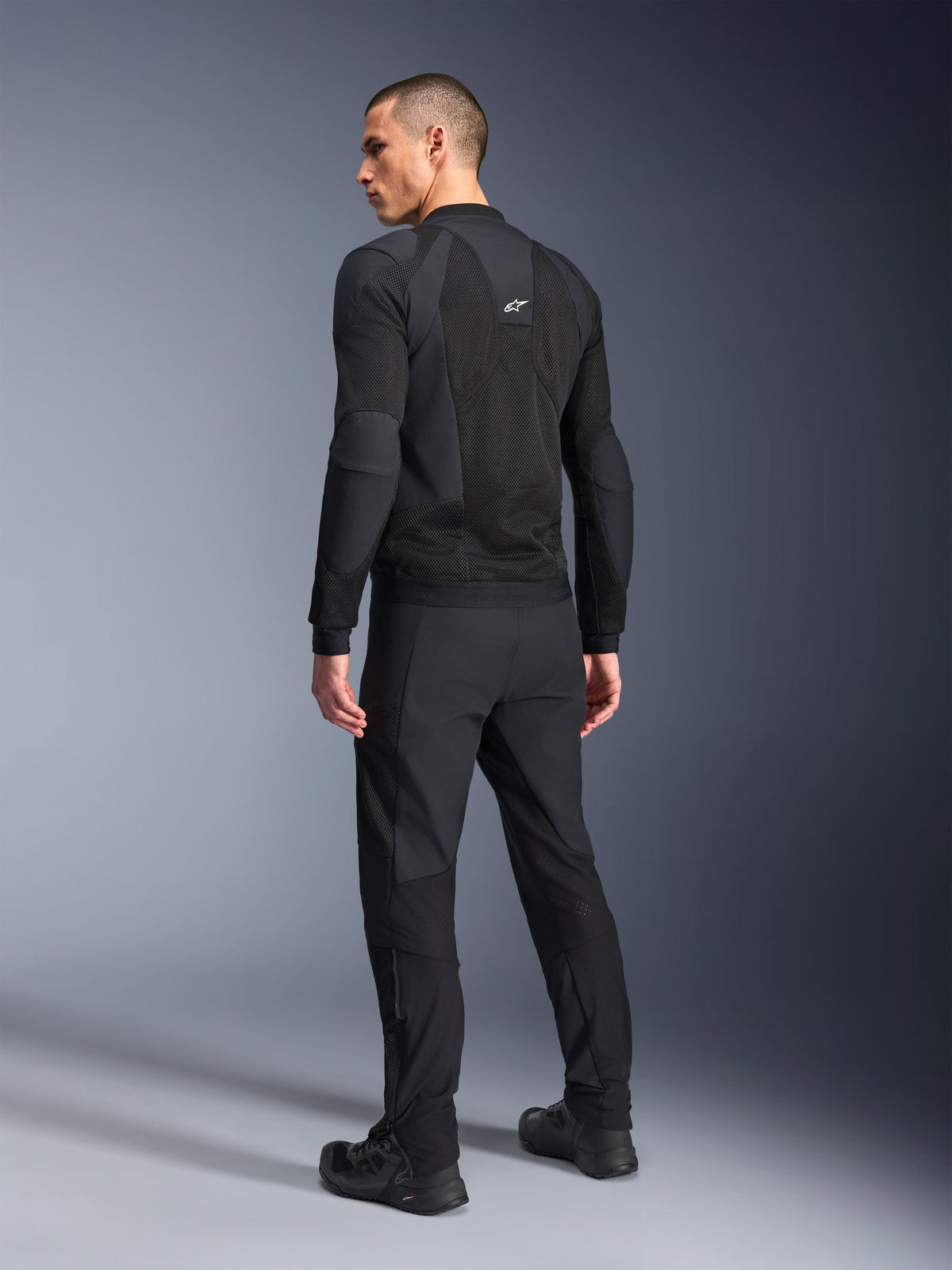 Alpinestars Troop-Air Pro Jacket, czarna, techniczna kurtka motocyklowa z pancerzem pokazana od tyłu, z rozległymi panelami z siatki zapewniającymi przepływ powietrza, w zestawie z dopasowanymi czarnymi spodniami technicznymi i butami.