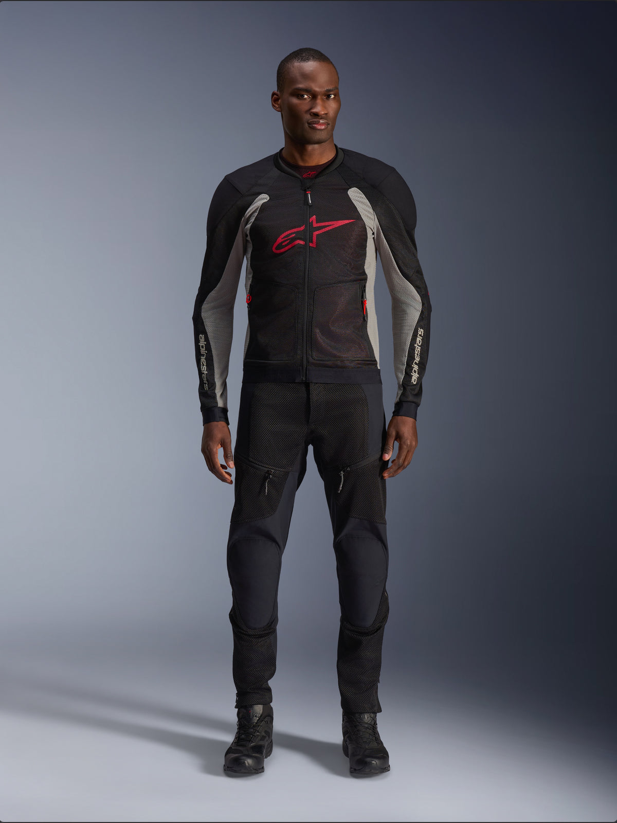 Męski model w kurtce Alpinestars Troop-Air w kolorze Black Ash Gray i Bright Red, techniczna siatkowa kurtka motocyklowa z wyraźnym czerwonym logo na klatce piersiowej, czarne spodnie i buty motocyklowe, miejska odzież motocyklowa