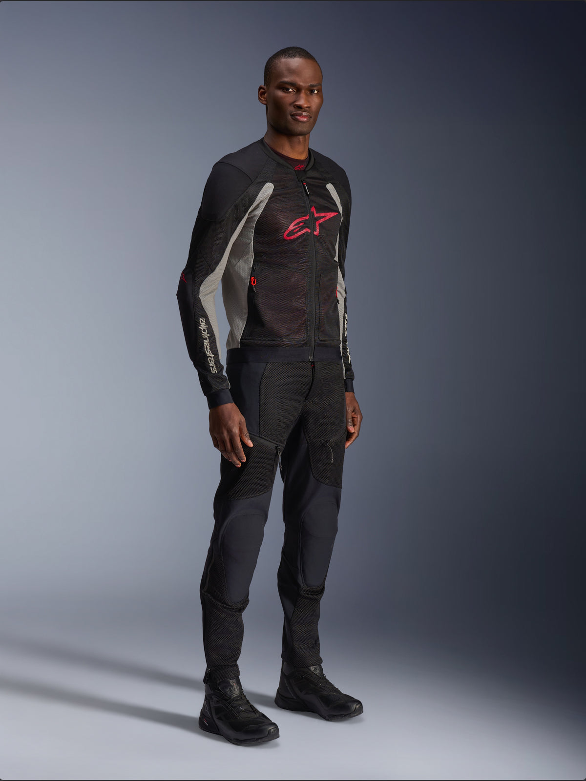 Kurtka Alpinestars Troop-Air, techniczna siatkowa kurtka motocyklowa w kolorach czarnym, popielatym i jasnoczerwonym, prezentowana przez modela z czarnymi spodniami i butami, czerwone logo Alpinestars na piersi, nowoczesna odzież motocyklowa w stylu miejskim.