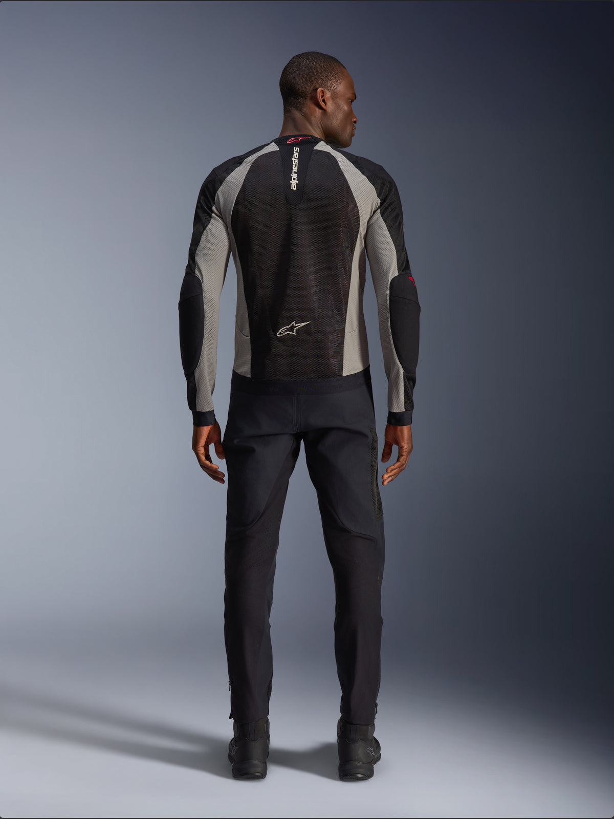 Alpinestars Troop-Air Jacket, motocyklowa kurtka w kolorze czarnym, popielatym i jaskrawoczerwonym, widok z tyłu na modelu, lekka siatkowa konstrukcja zapewniająca wentylację, branding Alpinestars na kołnierzu i w dolnej części pleców, w zestawieniu z czarnymi spodniami motocyklowymi