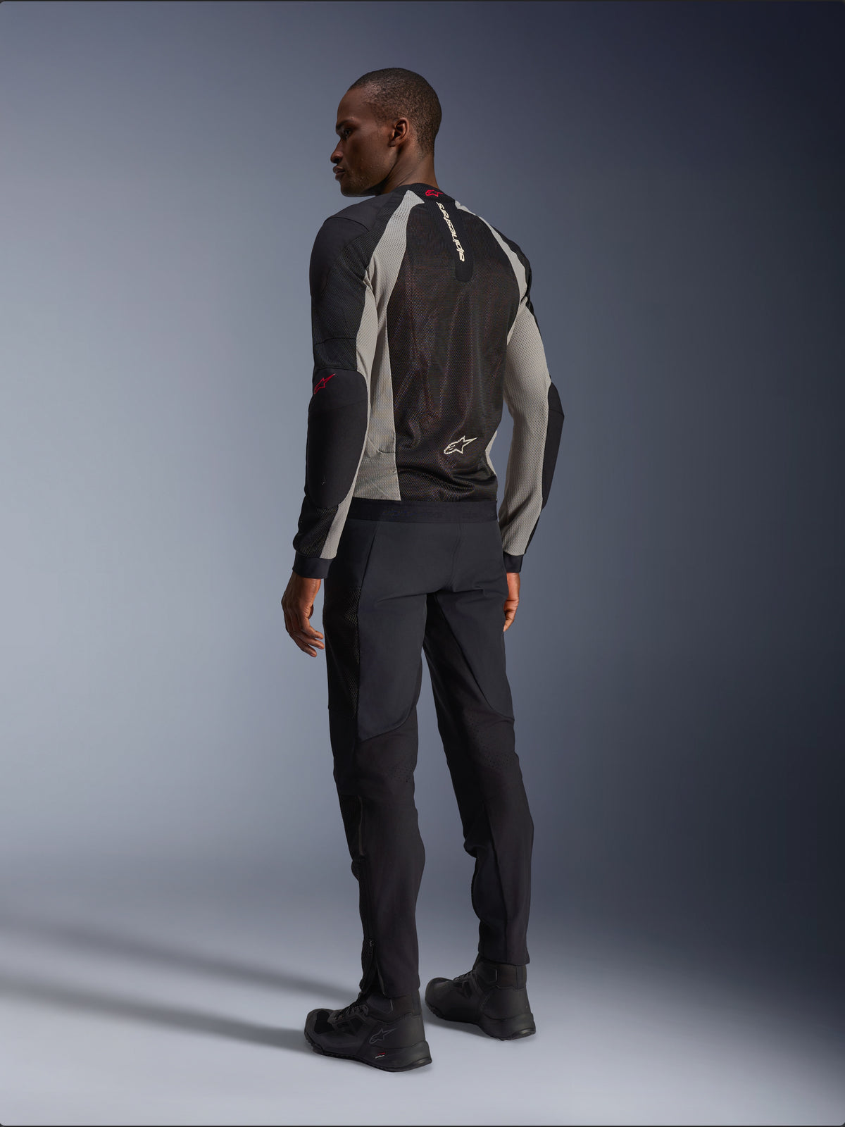 Model w kurtce Alpinestars Troop-Air, widok od tyłu, kolorystyka Black Ash Gray z jaskrawoczerwonymi akcentami, techniczna konstrukcja z siatki mesh dla optymalnej wentylacji, czarne spodnie motocyklowe i buty, współczesny styl miejski