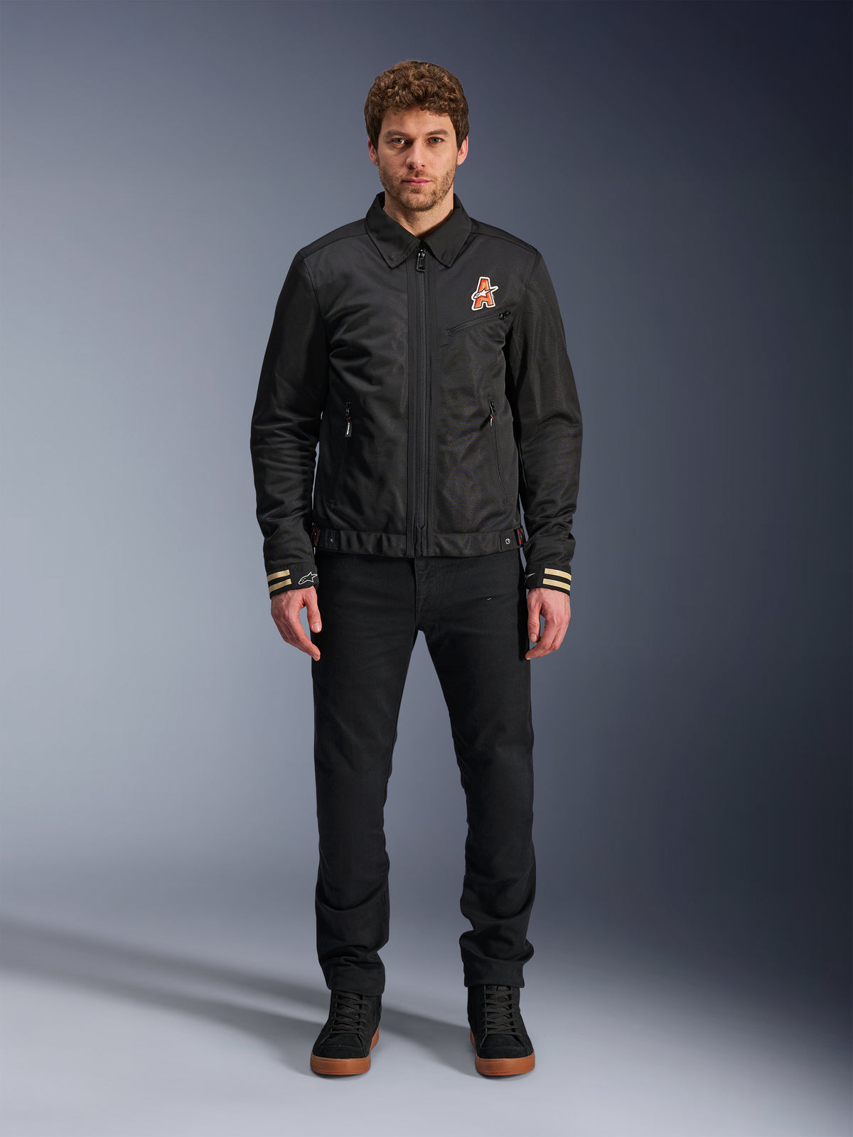 Męski model w czarnej kurtce Alpinestars Ignite Air z klasycznym kołnierzykiem koszulowym, pełnym zamkiem błyskawicznym z przodu i pomarańczowym logo na piersi. Stylizacja z czarnymi spodniami i czarnymi sneakersami za kostkę tworzy współczesny miejski wygląd motocyklowy.