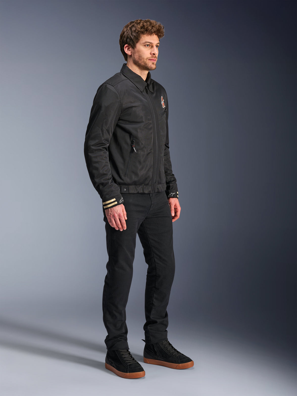 Alpinestars Ignite Air Jacket, miejska kurtka motocyklowa, czarna, prezentowana przez modela w czarnych spodniach i czarnych sneakersach, z tradycyjnym kołnierzykiem koszulowym, grafikami inspirowanymi stylem klubowym na klatce piersiowej i mankietach, elegancki, nowoczesny design z wykończeniem z siatki i satyny