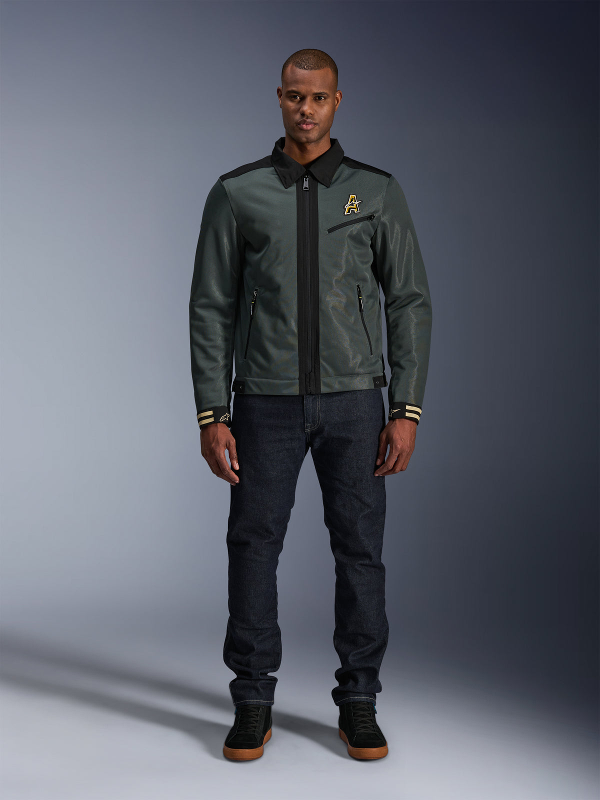 Kurtka Alpinestars Ignite Air w kolorze Urban Gray Black Blue, kurtka motocyklowa w stylu Harrington z kołnierzykiem koszulowym i siateczkową konstrukcją, noszona przez mężczyznę w ciemnych jeansach i czarnych sneakersach, miejski streetwear dla motocyklistów.