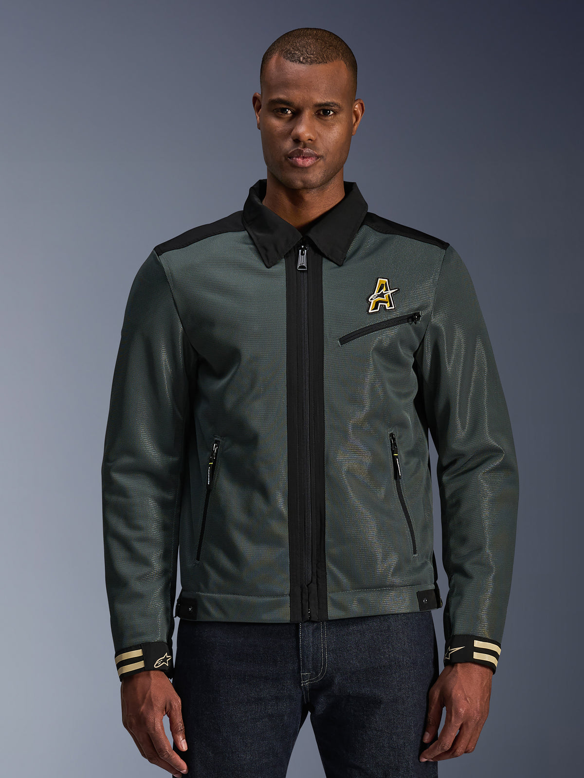Alpinestars Ignite Air Jacket, miejska kurtka motocyklowa, ciemnozielona z czarnymi akcentami, zaprezentowana na modelu w jeansach, kołnierzyk typu Harrington, zapięcie na zamek, logo na piersi i mankiety w paski, nowoczesna konstrukcja z siatki i materiału tekstylnego do jazdy latem