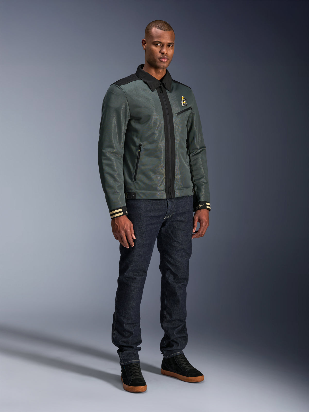 Alpinestars Ignite Air Jacket, Urban Gray Black Blue, miejska kurtka motocyklowa, noszona przez modela z ciemnymi jeansami i czarnymi sneakersami, siateczkowy korpus, tradycyjny kołnierzyk koszulowy, fason inspirowany kurtką Harrington i złote logo Alpinestars.
