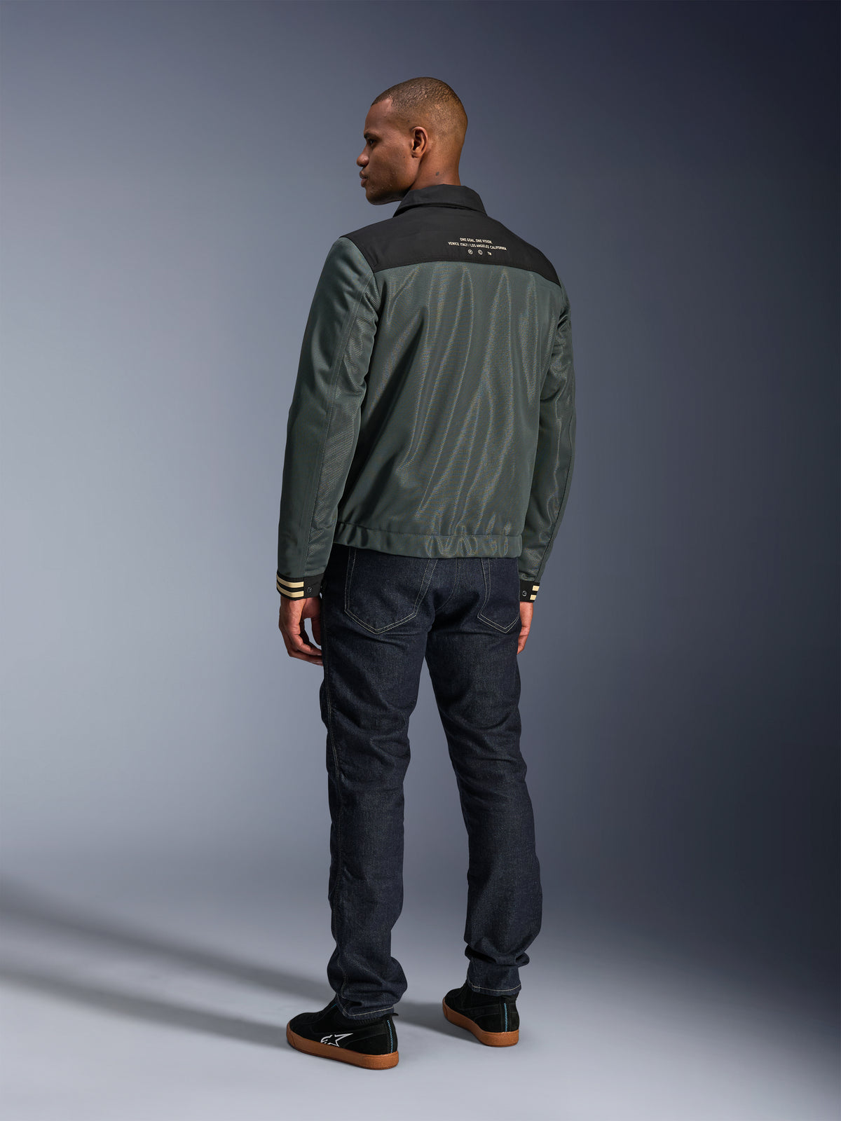 Kurtka Alpinestars Ignite Air w kolorze Urban Gray Black Blue, widok z tyłu na modelu, siatkowy korpus z czarnym karczkiem i białym napisem, prążkowane mankiety, w zestawieniu z ciemnymi jeansami i czarnymi sneakersami, miejska odzież motocyklowa lifestyle