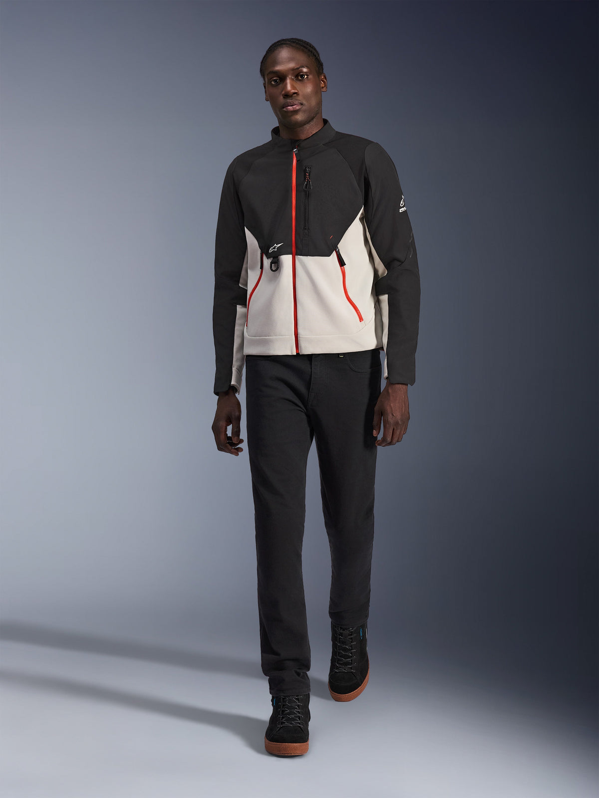 Męski model w kurtce Alpinestars Provoke, miejska kurtka motocyklowa w kolorze czarnym i metalicznym szarym z czerwonymi akcentami przy zamkach, zestawiona z czarnymi spodniami i czarnymi butami za kostkę, nowoczesna odzież ochronna do jazdy miejskiej