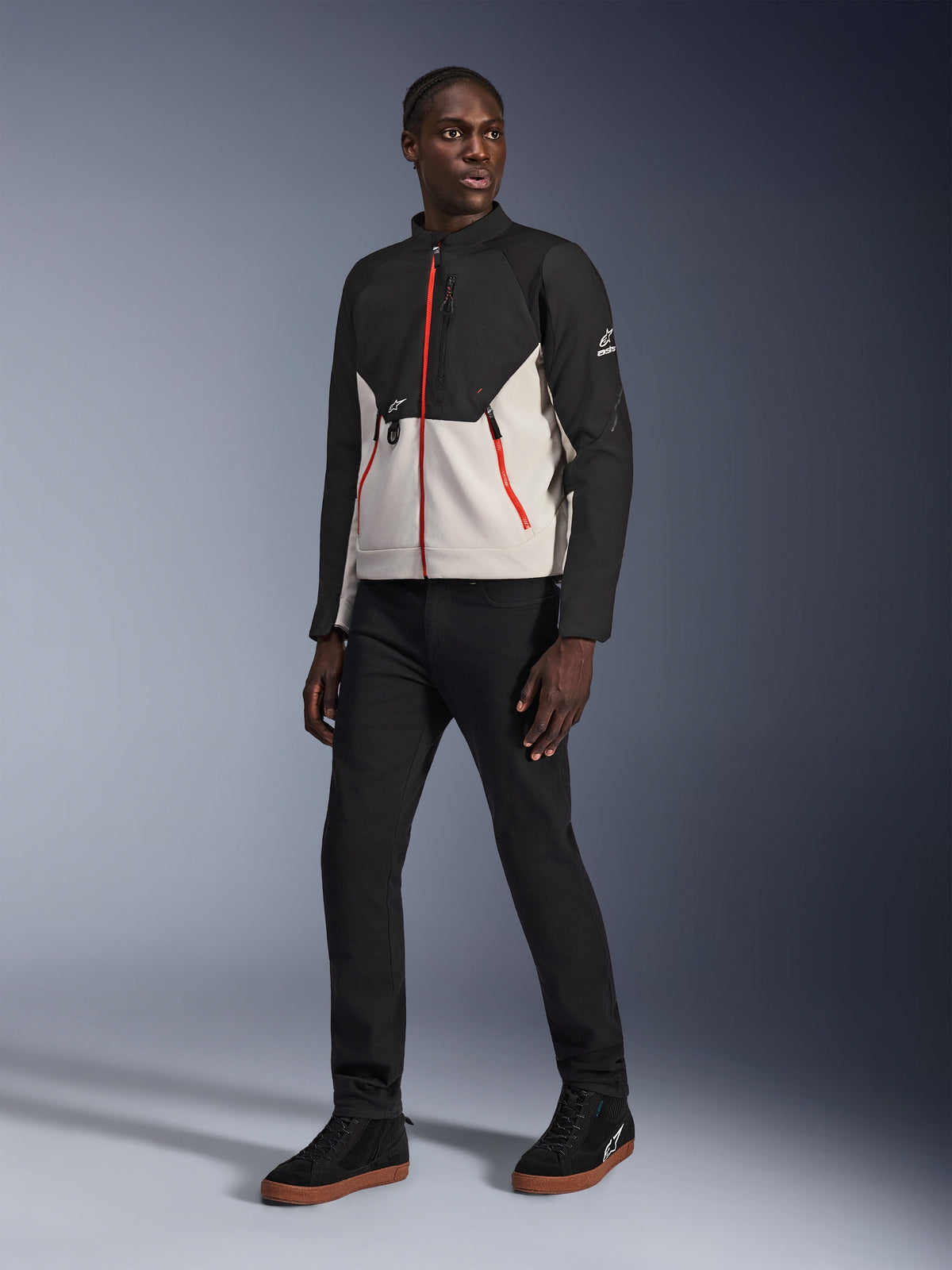 Model ubrany w kurtkę Alpinestars Provoke w kolorze Black Metal Gray z charakterystycznymi czerwonymi zamkami z przodu i przy kieszeniach, czarne spodnie oraz czarne wysokie sneakersy z gumową podeszwą; współczesna miejska odzież motocyklowa w ulicznym stylu.