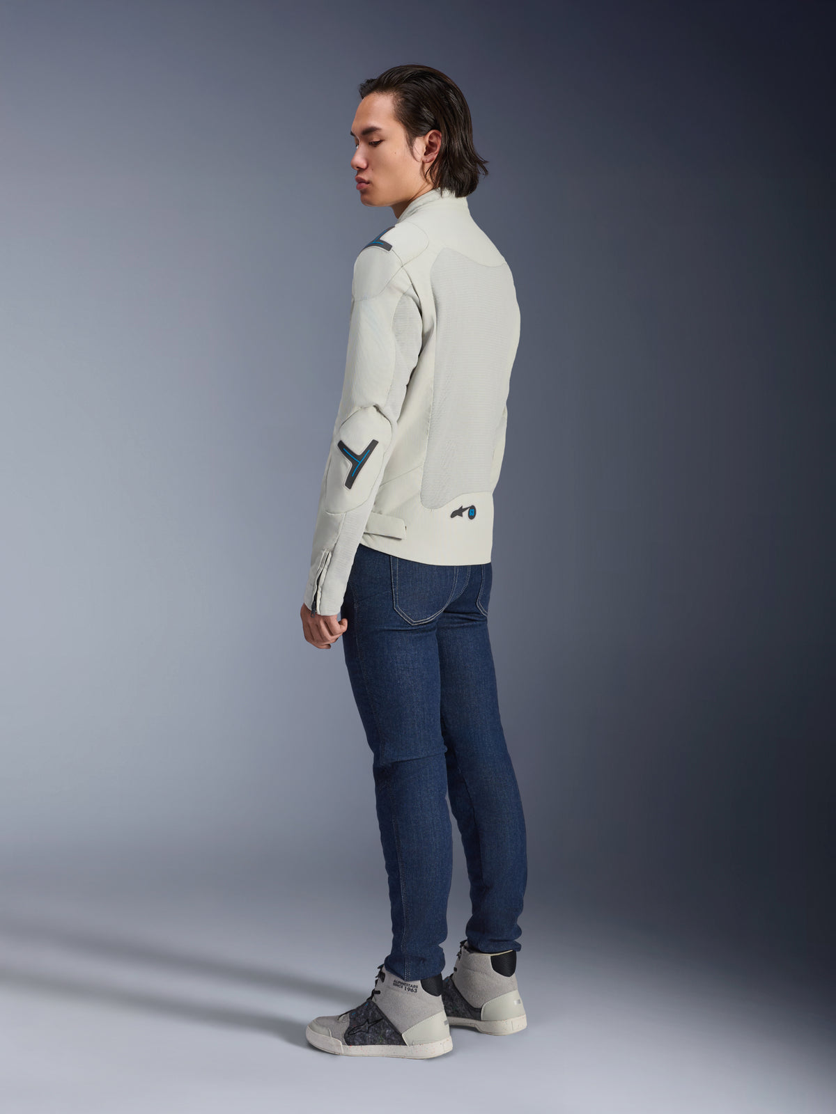 Kurtka Alpinestars Circal-1 Air, miejska kurtka motocyklowa, kolor Metal Gray z akcentami Methyl Blue, noszona przez modela widocznego z tyłu pod kątem, w ciemnoniebieskich dżinsach i szarych wysokich tenisówkach, panele siatkowe i minimalistyczny design