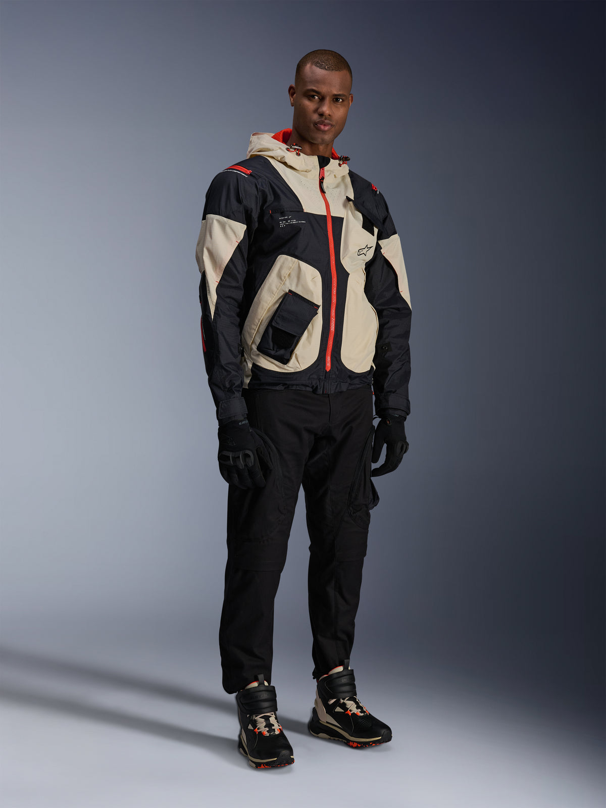 Mężczyzna w pełnym miejskim rynsztunku motocyklowym, buty Alpinestars Speedstreet w kolorze Black Almond Hot Orange, kurtka Mogress Air w kolorze czarnym i kremowym, dopasowane czarne spodnie i rękawice, ochronny strój inspirowany stylem streetwear do jazdy miejskiej