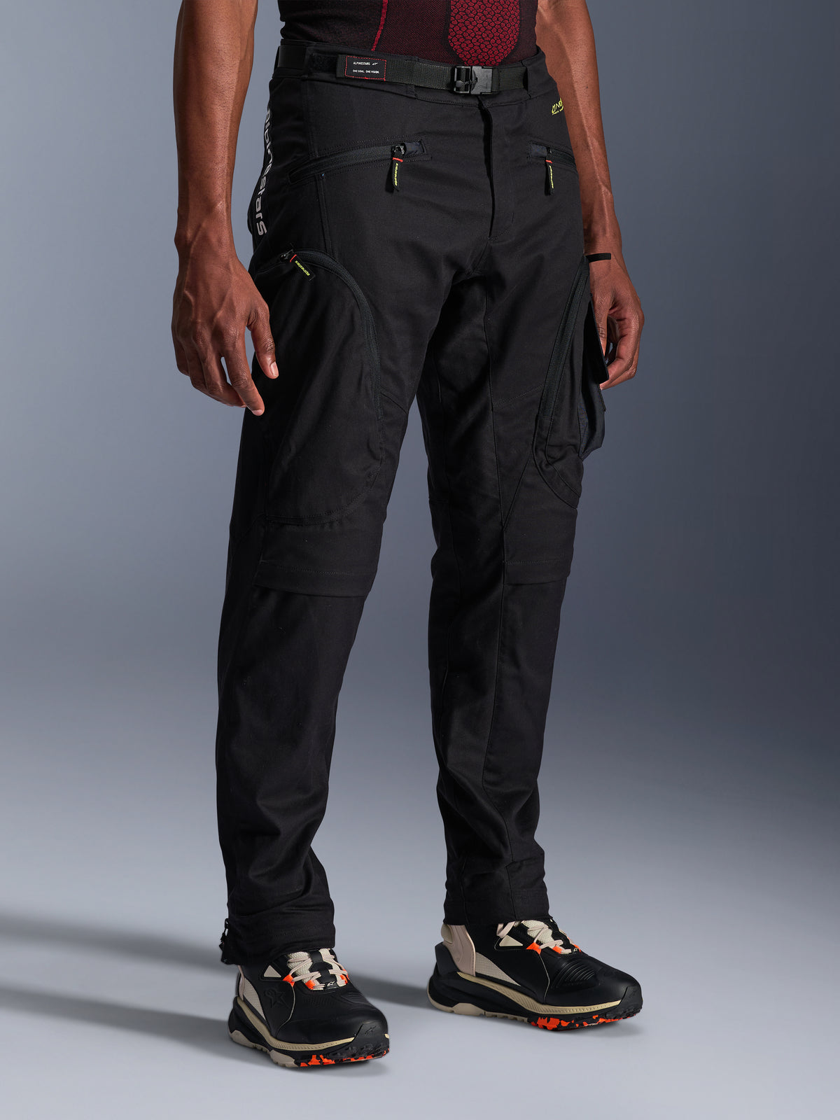 Alpinestars Acteon Pants, czarne spodnie motocyklowe prezentowane na modelu, o nowoczesnym, zwężanym kroju, z kieszeniami cargo zapinanymi na zamek, regulowanym paskiem w talii i technicznymi sneakersami, miejska odzież motocyklowa
