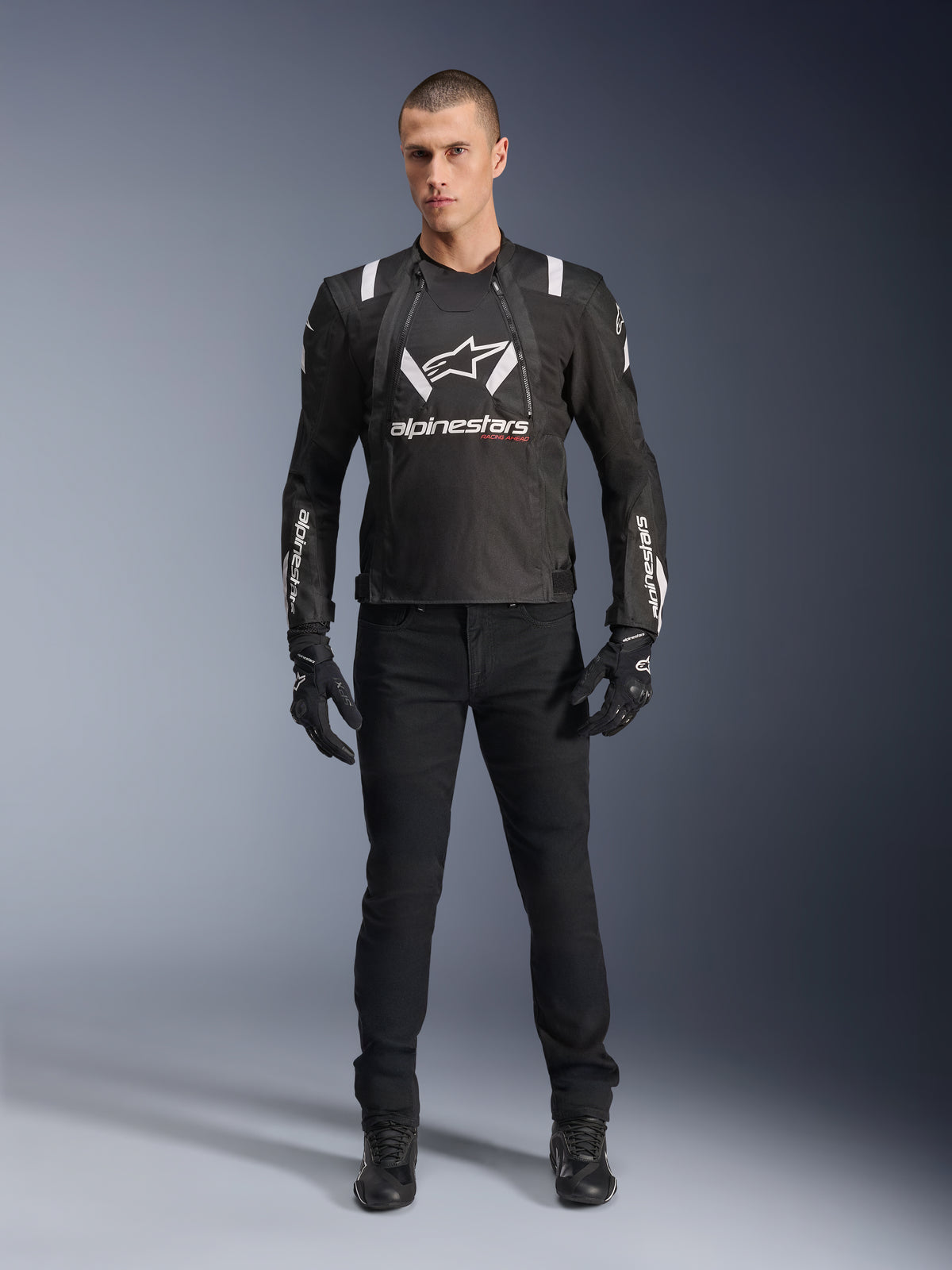 Kurtka Alpinestars T-Stunt Air, czarno-biała kurtka motocyklowa, noszona z czarnymi spodniami motocyklowymi, rękawicami i butami, z wyraźnym białym logo Alpinestars na klatce piersiowej i rękawach, sportowy krój tekstylny z panelami z siatki dla wentylacji