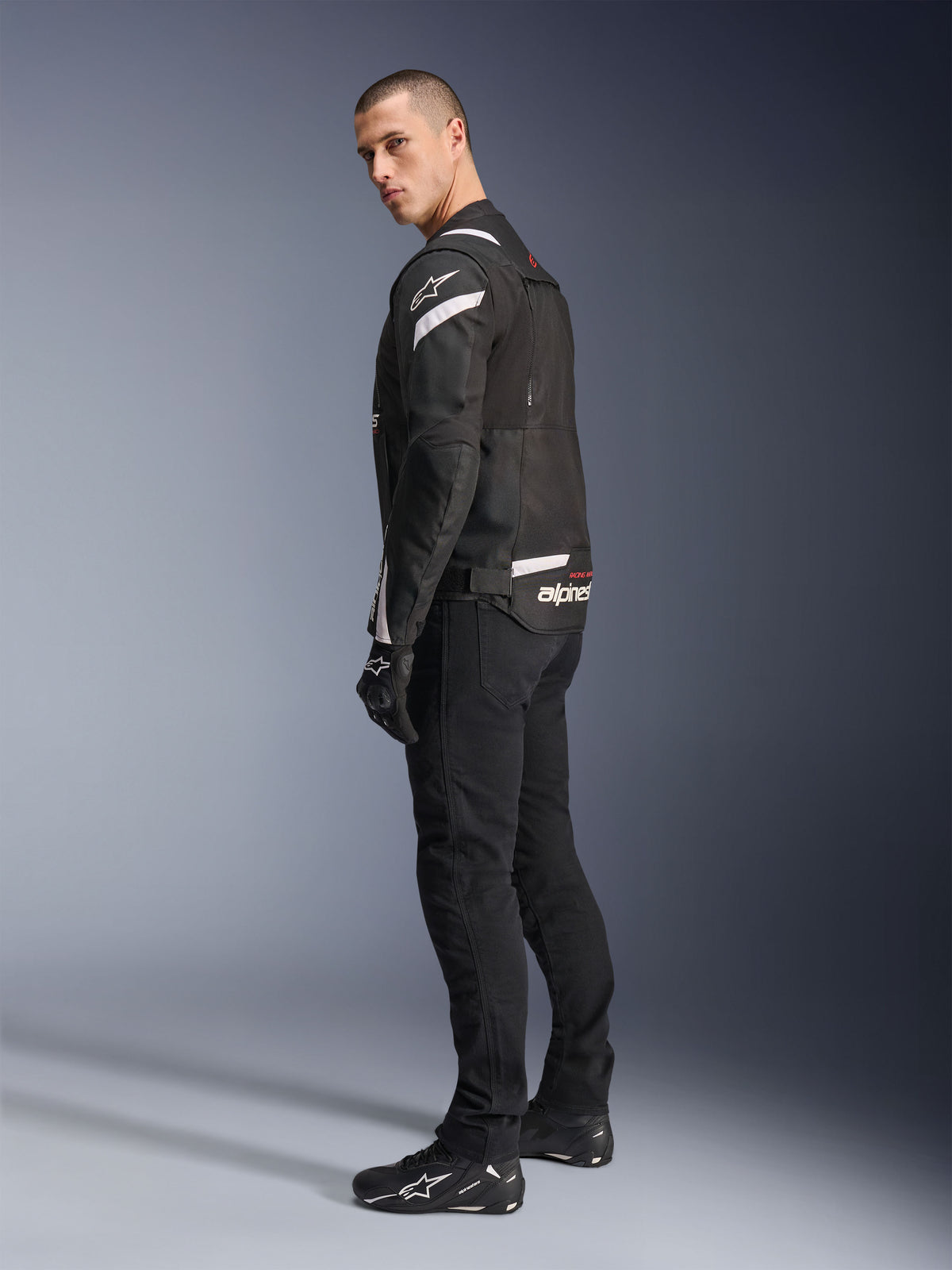 Alpinestars T-Stunt Air Jacket, czarno-biała, modułowa kurtka motocyklowa z panelami z siatki technicznej, pokazana z profilu od tyłu na modelu, noszona z czarnymi spodniami i butami motocyklowymi, z białymi akcentami i logo na ramionach i plecach.