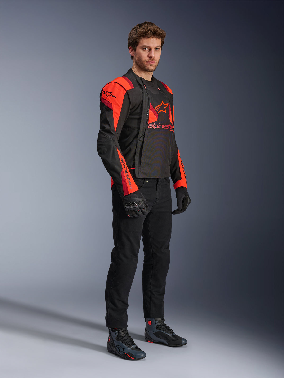 Alpinestars T-Stunt Air Jacket, czarna z elementami czerwonymi fluo i ciemnoczerwonymi, modułowa kurtka motocyklowa prezentowana przez modela z otwartymi panelami przednimi ukazującymi siatkowe wnętrze, zestawiona z czarnymi spodniami, rękawicami i butami motocyklowymi