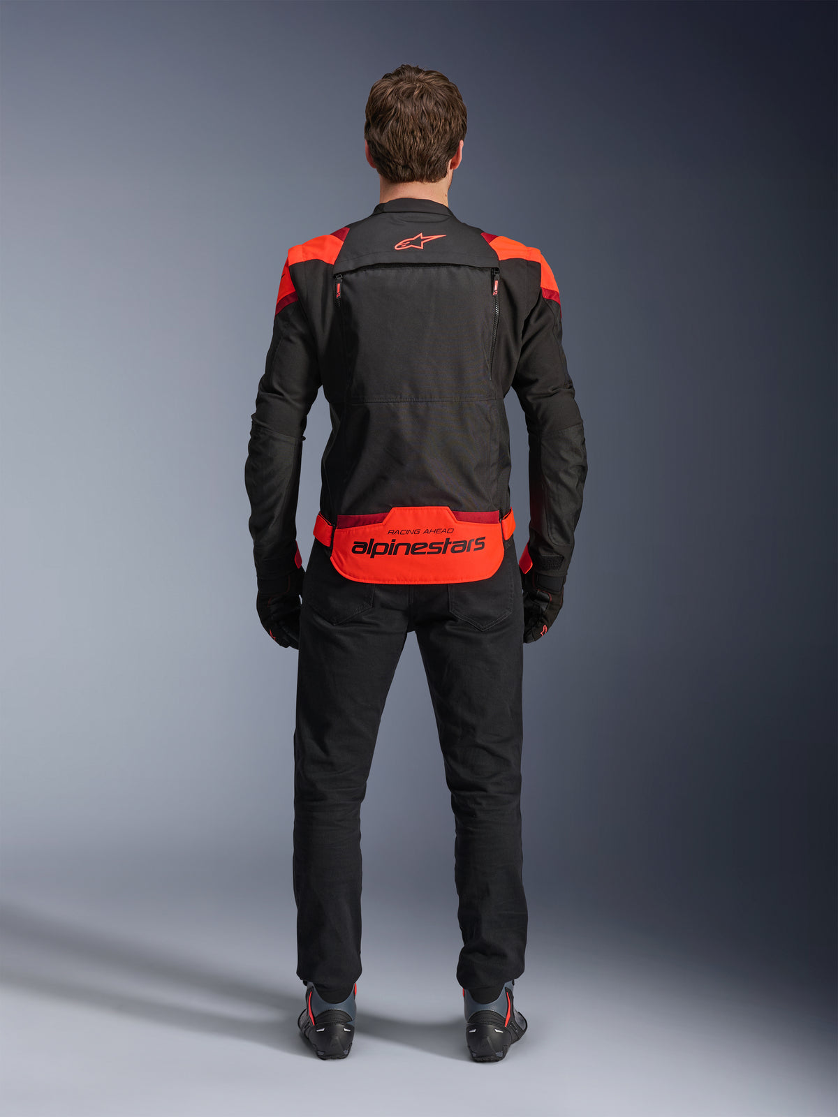 Kurtka Alpinestars T-Stunt Air, czarna z akcentami Red Fluo i ciemnoczerwonymi, widok z tyłu na modelu, w zestawieniu z czarnymi spodniami i butami motocyklowymi, wyraźne logo Alpinestars na dolnym panelu pleców, modułowa konstrukcja z siateczki zapewniająca wentylację