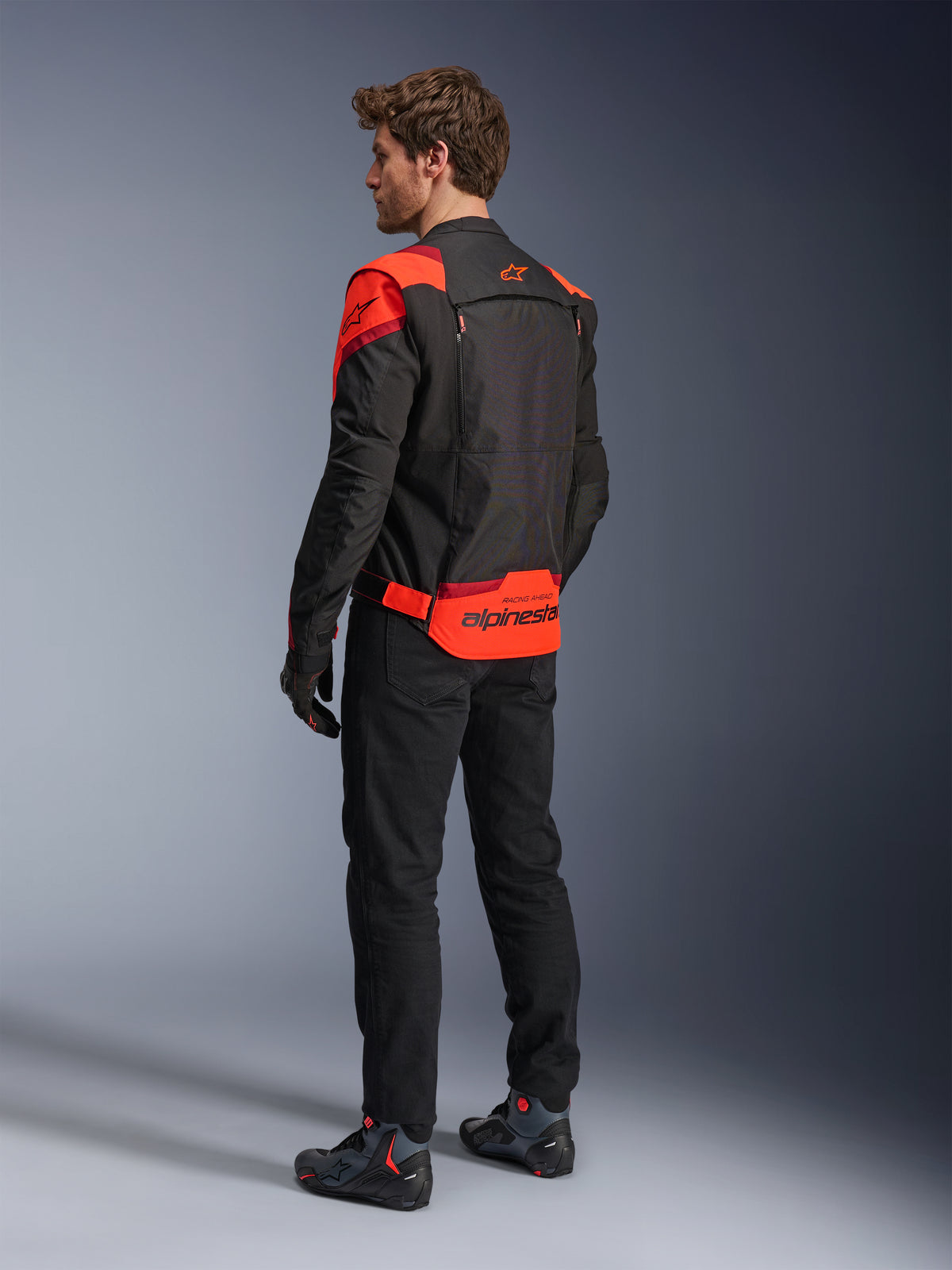Mężczyzna w kurtce Alpinestars T-Stunt Air Jacket, widok z tyłu, kolor czarny, neonowy czerwony i ciemnoczerwony, tekstylna kurtka motocyklowa z panelami z siatki o sportowym kroju, z wyraźnym brandingiem Alpinestars na dole pleców, zestawiona z czarnymi spodniami i butami motocyklowymi