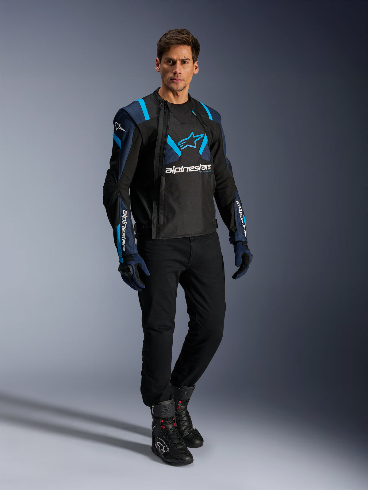 Męski model w kurtce Alpinestars T-Stunt Air, kolor czarny, granatowy i błękitny (Blithe Blue), z widocznymi panelami z siatki dla lepszego przepływu powietrza, w zestawieniu z czarnymi spodniami i rękawicami motocyklowymi, sportowa kurtka motocyklowa do letniej jazdy szosowej.