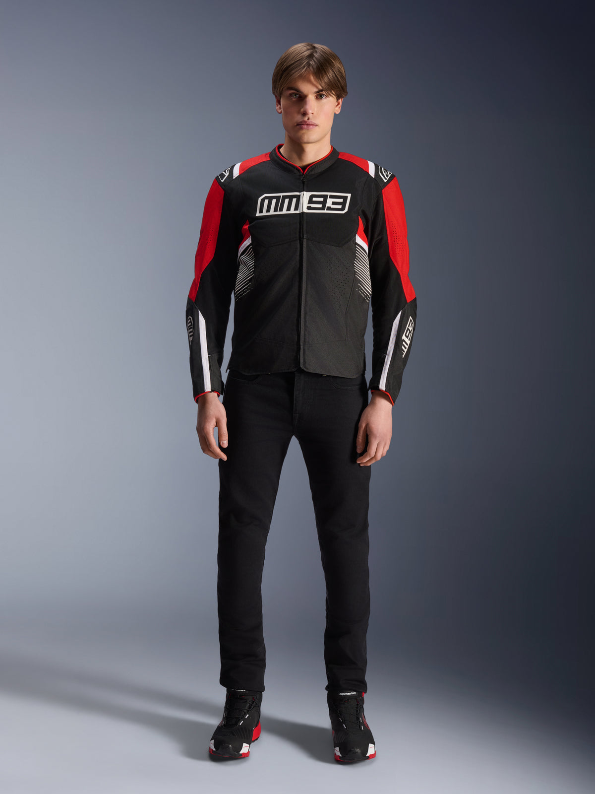 Alpinestars MM93 T-SPS Air V2 Jacket, męska kurtka motocyklowa, czarna z jasnoczerwonymi i białymi detalami, logo Marca Márqueza MM93 na piersi, panele z siatki dla przepływu powietrza, model w czarnych spodniach i butach sportowych w studio.