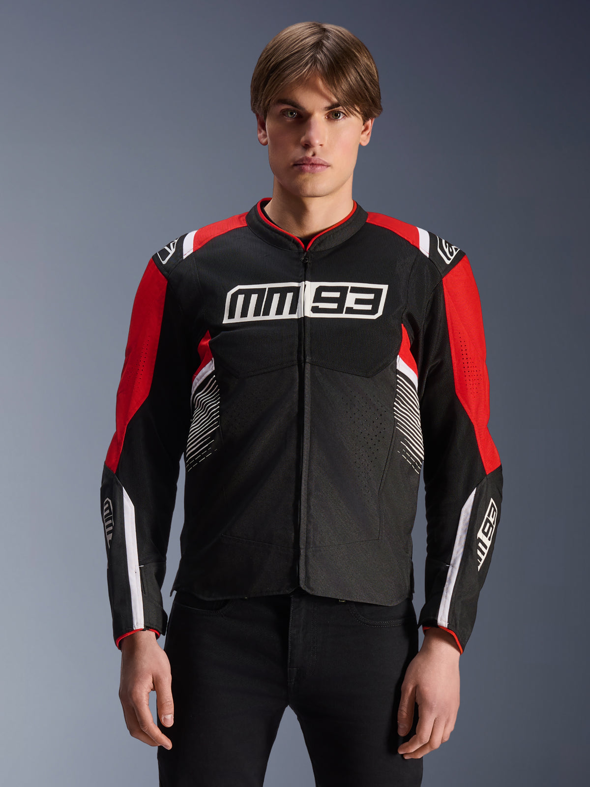 Kurtka MM93 T-SPS Air V2, tekstylna kurtka motocyklowa w kolorze czarnym, jaskrawoczerwonym i białym, z logo MM93 Marca Márqueza na klatce piersiowej i rękawach, panele z siatki dla wentylacji, sportowy krój, prezentowana przez modela w czarnych spodniach