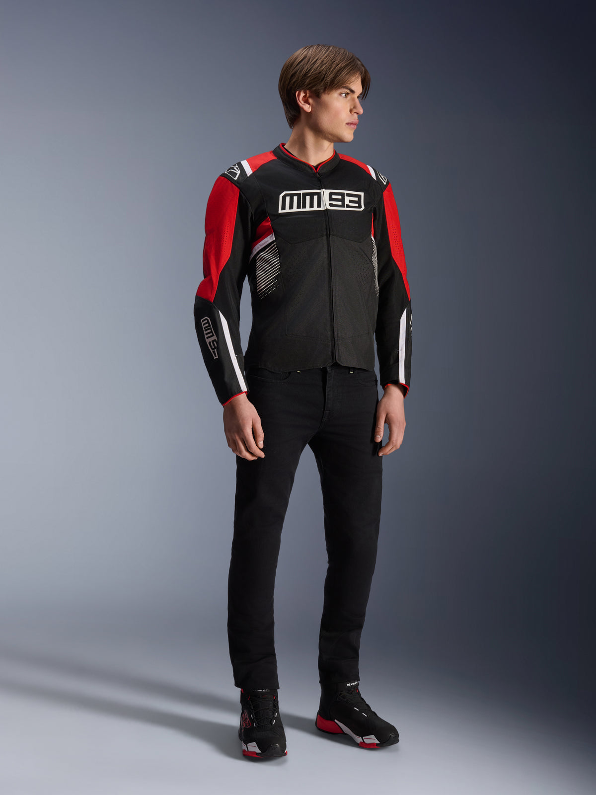Męski model ubrany w kurtkę Alpinestars MM93 T-SPS Air v2 w kolorze Black Bright Red White, z wyraźnym logo MM93 na klatce piersiowej i rękawach, w zestawieniu z czarnymi spodniami i butami motocyklowymi, sportowy miejski styl motocyklowy z widocznymi panelami z siatki zapewniającymi przepływ powietrza.