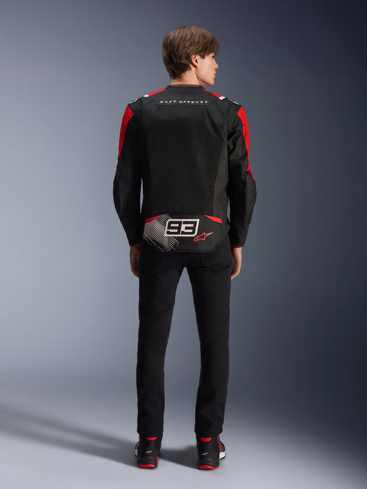 Kurtka Alpinestars MM93 T-SPS Air V2, widok z tyłu na modelu, czarna z jaskrawoczerwonymi i białymi akcentami, z brandingiem Marca Marqueza i logo 93 na plecach, tekstylna kurtka motocyklowa do jazdy miejskiej