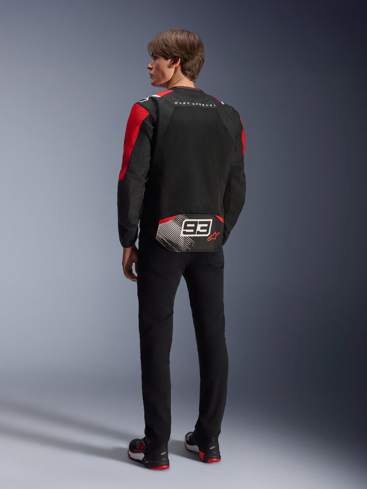 Model widziany od tyłu w kurtce Alpinestars MM93 T-SPS Air V2, czarna z jaskrawoczerwonymi i białymi detalami, napis „MARC MARQUEZ” i duże logo „93” na dole pleców, tekstylna konstrukcja z siatki zapewniająca wentylację, czarne spodnie i dopasowane czarno-czerwone buty motocyklowe