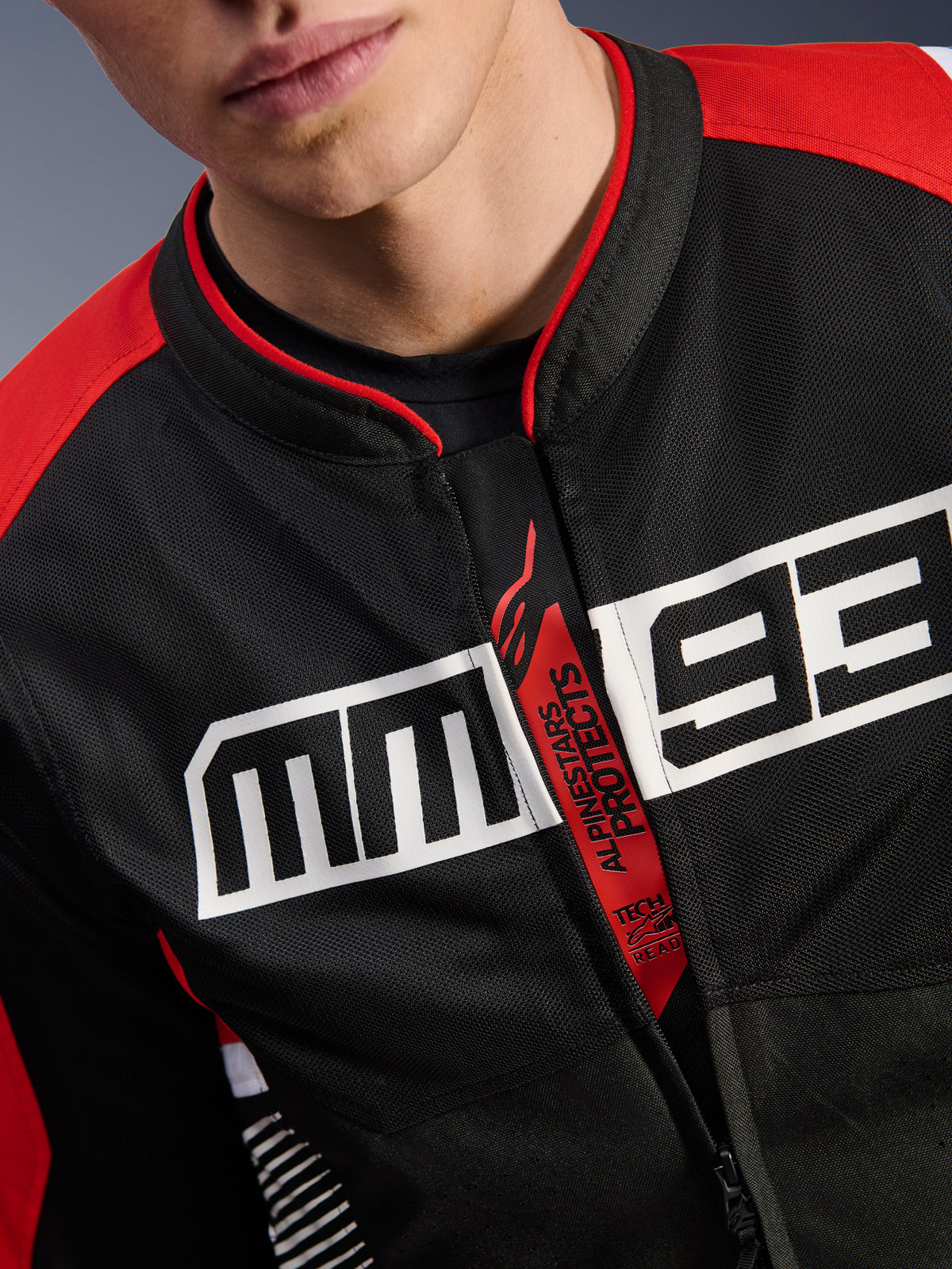 Kurtka Alpinestars MM93 T-SPS Air V2, kurtka motocyklowa, czarna, jaskrawoczerwona i biała, zbliżenie na klatkę piersiową z dużym białym logo MM93, czerwony uchwyt zamka z napisami „Alpinestars Protects” i „Tech-Air Ready”, tekstura materiału z technicznej siatki