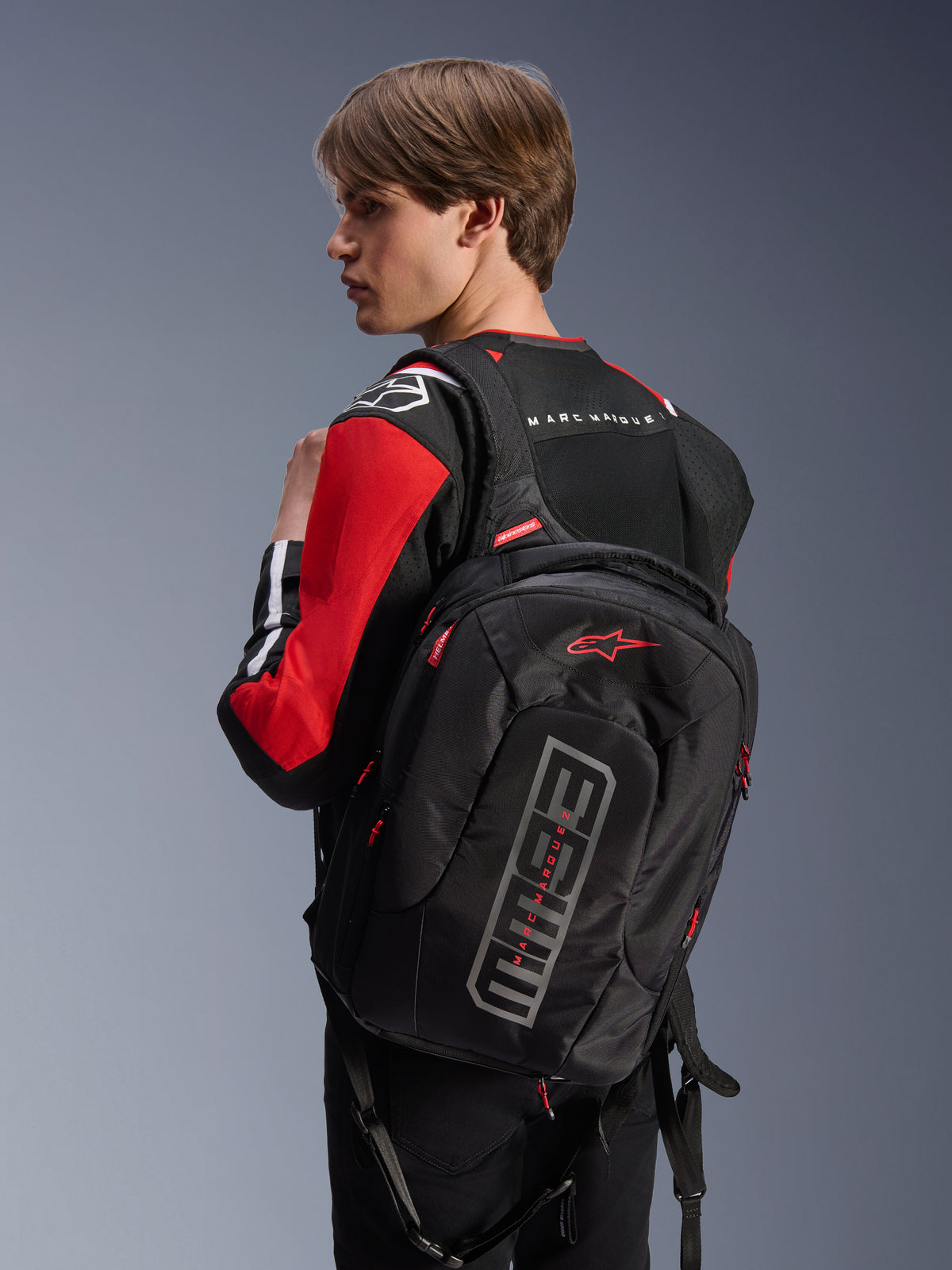 Model w kurtce Alpinestars MM93 T-SPS Air V2 w kolorze czarnym, jaskrawoczerwonym i białym, widok z tyłu z czarnym plecakiem Alpinestars z logo Marc Márquez, miejska odzież motocyklowa z panelami z siatki i detalami z logo