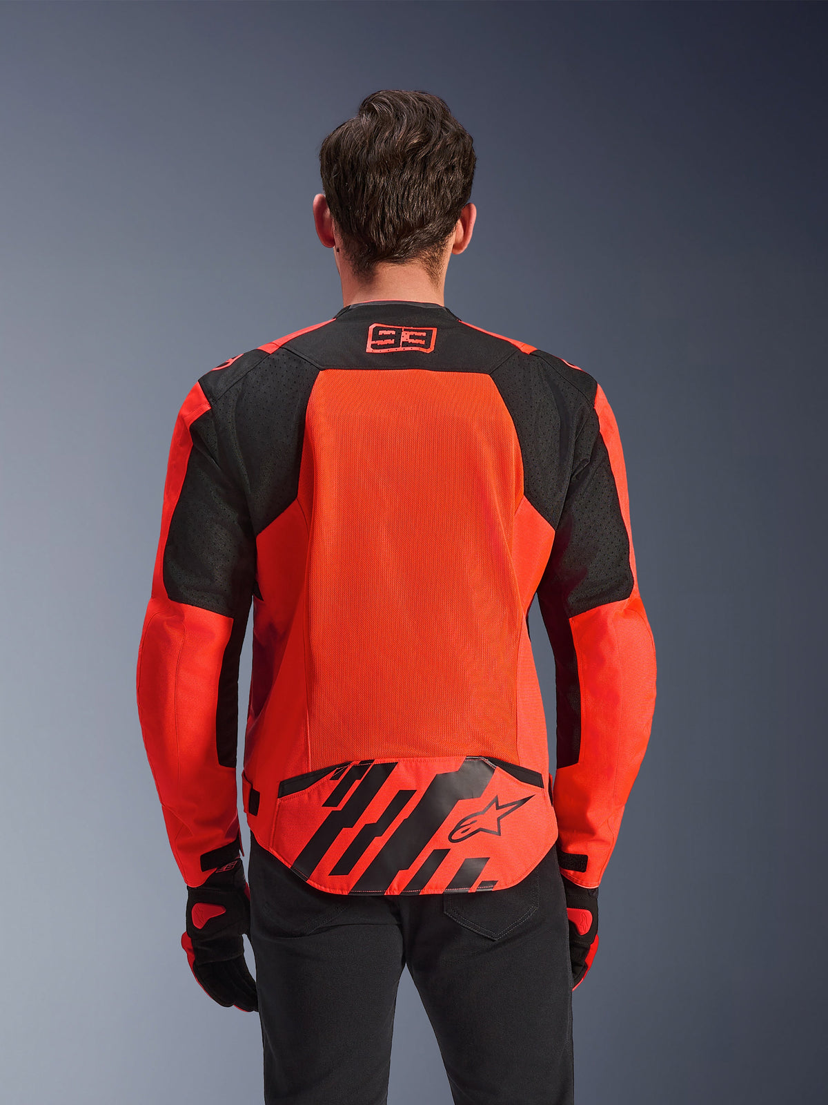 Kurtka Alpinestars MM93 T-SPS Air V2, widok z tyłu, kolor czerwony fluo i czarny, logo wyścigowe 93 na karku, duży panel z siatki na plecach dla optymalnego przepływu powietrza, dolna patka z logo gwiazdy Alpinestars i geometrycznymi pasami, noszona z czarnymi spodniami i rękawicami motocyklowymi