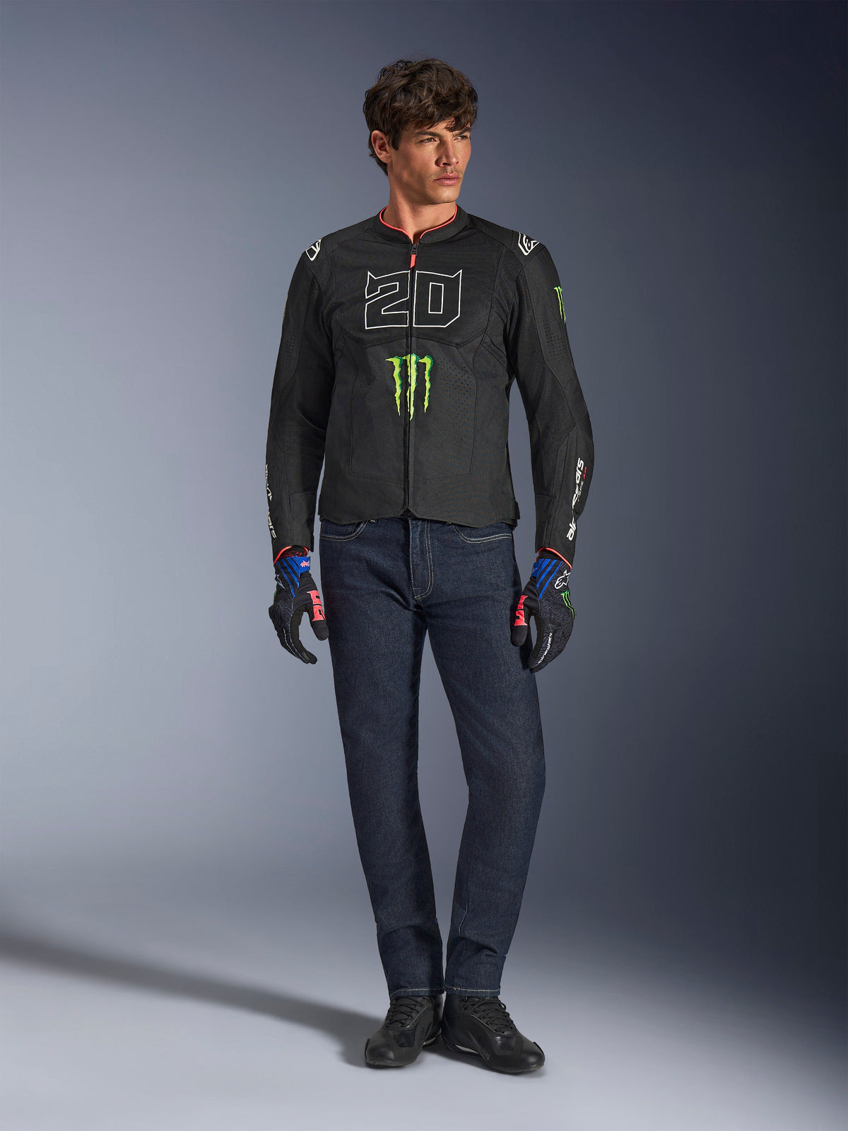 Male rider wearing the Alpinestars FQ20 T-SPS Air V2 Monster Jacket, black with green Monster Energy logo and white '20' graphic on the chest, paired with dark blue jeans and technical riding gloves, urban sport motorcycle outfit.