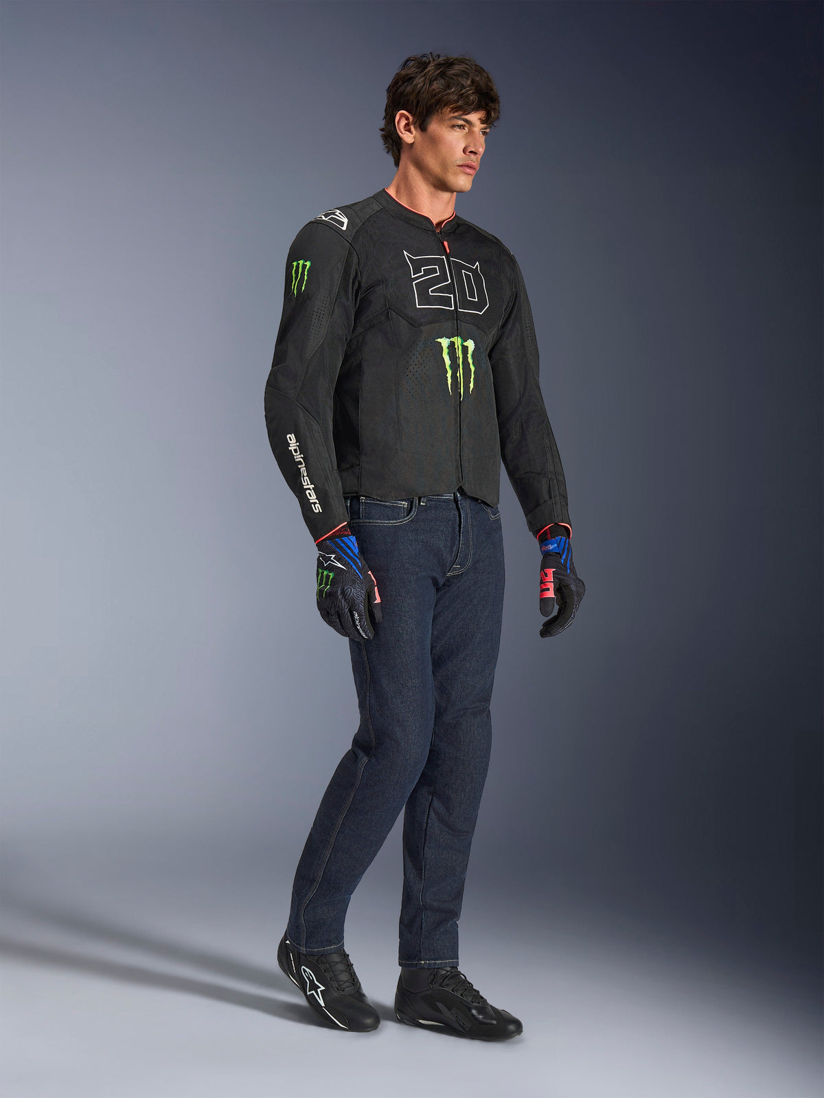Male rider wearing the Alpinestars FQ20 T-SPS Air V2 Monster Jacket in black, featuring the '20' logo and Monster Energy claw on the chest, paired with dark riding jeans, black motorcycle shoes, and matching FQ20 gloves, full-body studio shot.