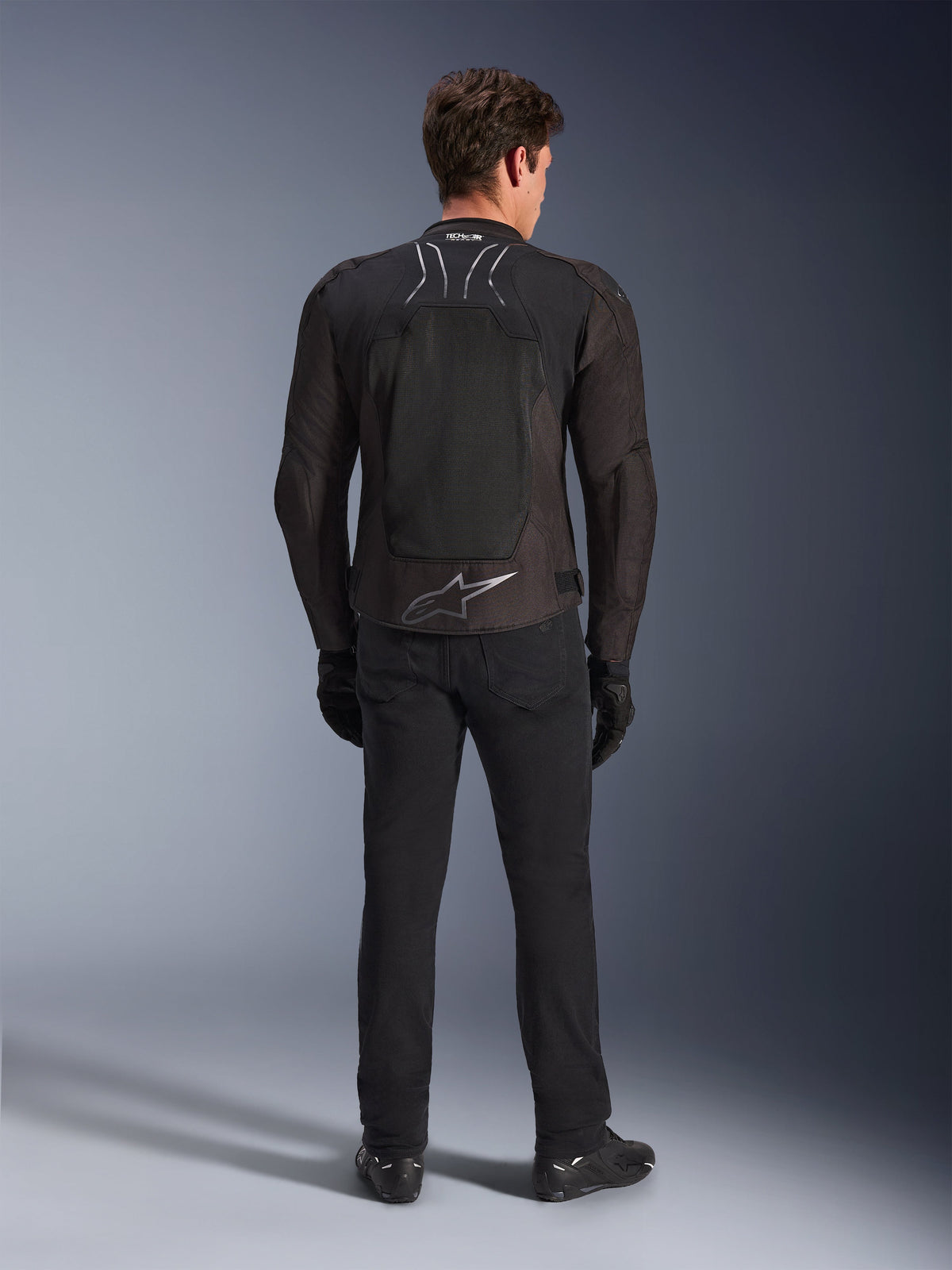 Alpinestars SMX Air Jacket, Black Anthracite, widok z tyłu na modelu, z dużym srebrnym logo Alpinestars w dolnej części pleców i panelami z siatki o wysokim przepływie powietrza, noszona z czarnymi spodniami motocyklowymi, rękawicami i butami, sportowa odzież motocyklowa