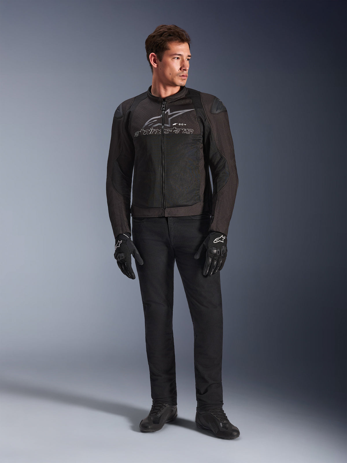 Męski model w kurtce motocyklowej Alpinestars SMX Air, kolor Black Anthracite, wentylowane panele z siatki i szare logo na klatce piersiowej, czarne spodnie i rękawice ochronne, tło studyjne z gradientem