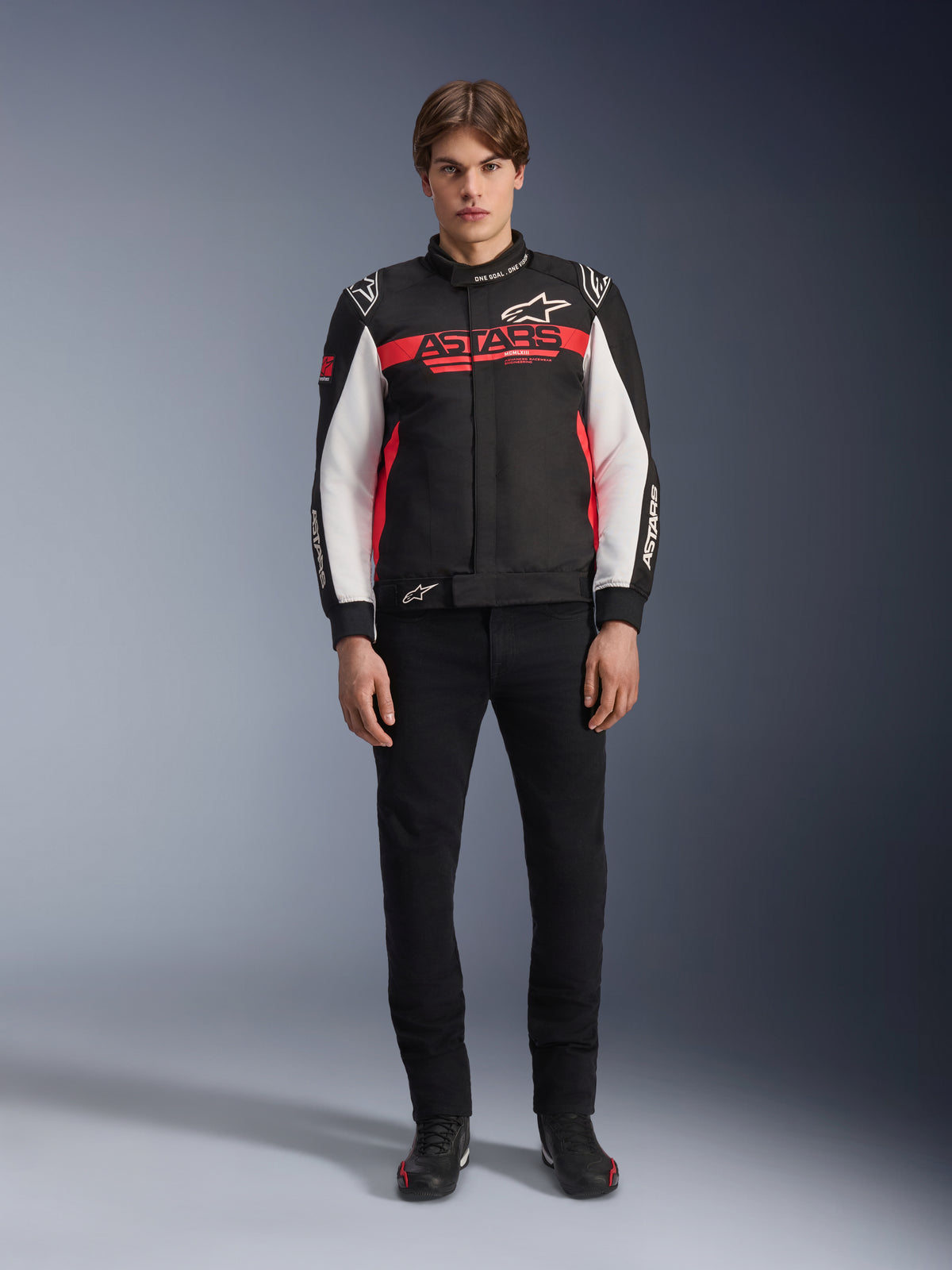 Męski model w kurtce Alpinestars Monza Sport w kolorze czarnym, jaskrawoczerwonym i białym, z dużym czerwonym logo „ASTARS” na klatce piersiowej i białymi panelami na rękawach, w zestawieniu z czarnymi spodniami i czarnymi butami motocyklowymi.