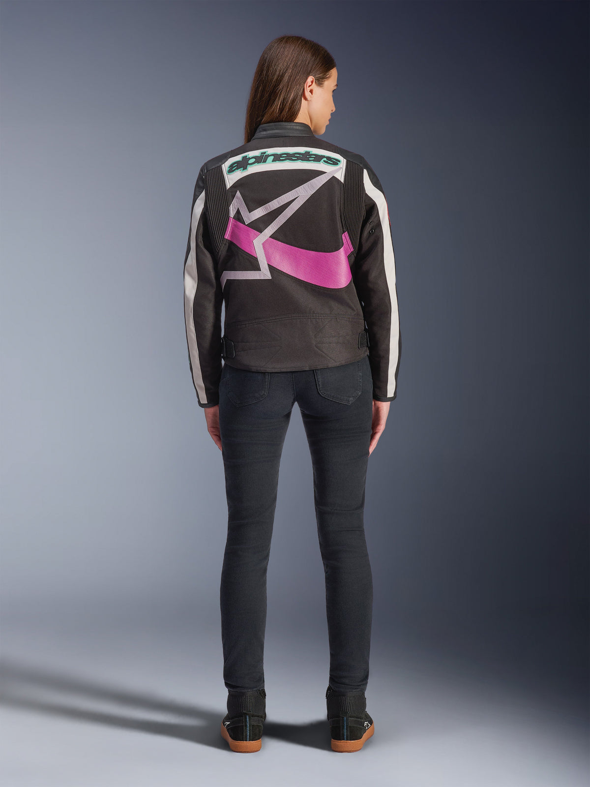 Widok z tyłu kobiety w kurtce Alpinestars Idles, czarna z pasami w kolorze złamanej bieli, z dużym fioletowo-białym logo gwiazdy Alpinestars i zielonym napisem na plecach, zestawiona z ciemnymi dopasowanymi jeansami, miejska odzież motocyklowa w stylu vintage