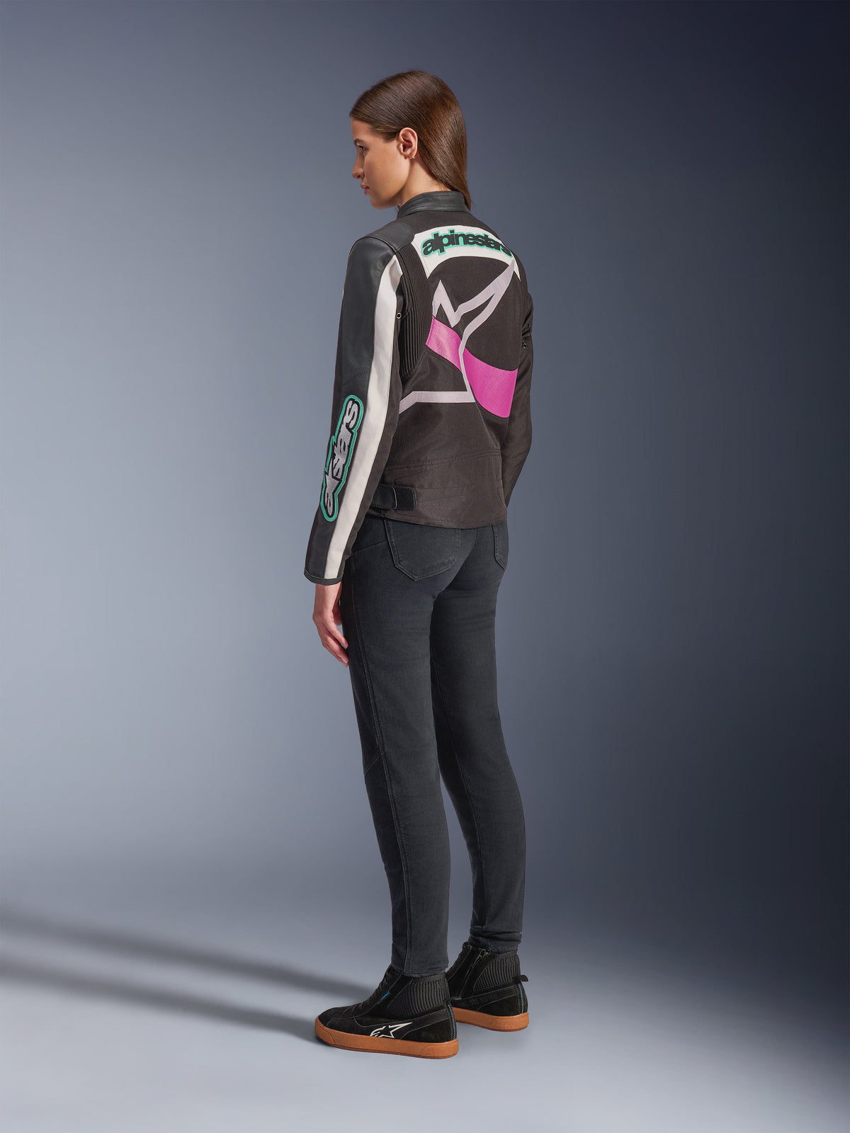 Damska kurtka motocyklowa Alpinestars Idles, czarna, kremowa i fioletowa, widok z tyłu na modelce, z dużym, oplatającym logo Alpinestars i designem inspirowanym stylem vintage racing, zestawiona z ciemnymi dżinsami i czarnymi butami.