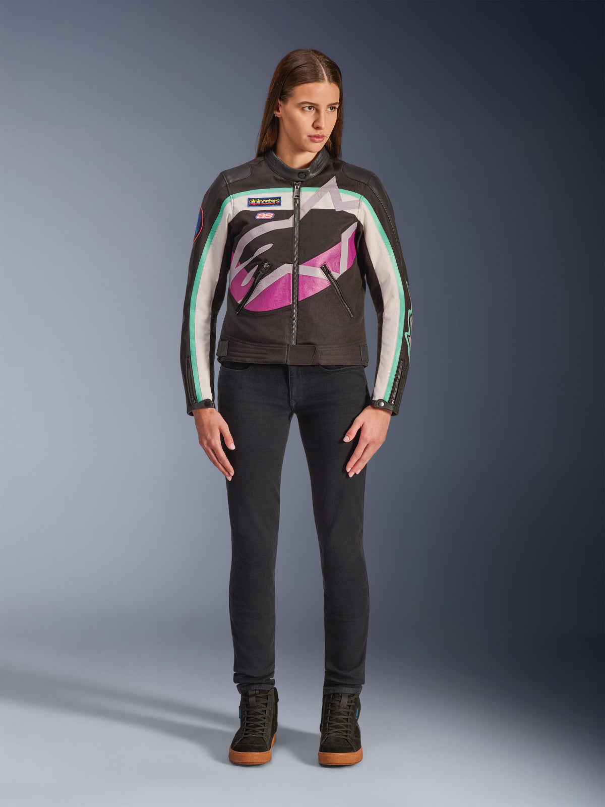 Kobieta ubrana w damską kurtkę Alpinestars Idles, czarna ze złamaną bielą i fioletem, wzór inspirowany wyścigami vintage z dużym otaczającym logo i turkusowymi akcentami, stylizacja z czarnymi jeansami i butami motocyklowymi, miejska odzież motocyklowa