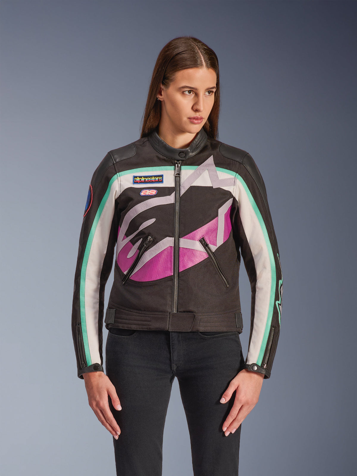 Damska kurtka Alpinestars Idles, kurtka motocyklowa, czarna z biało-fioletowo-turkusowym wzorem blokowym, styl inspirowany wyścigami vintage z dużym abstrakcyjnym logo na piersi, markowe naszywki i zamek błyskawiczny z przodu, noszona przez modelkę z czarnymi jeansami