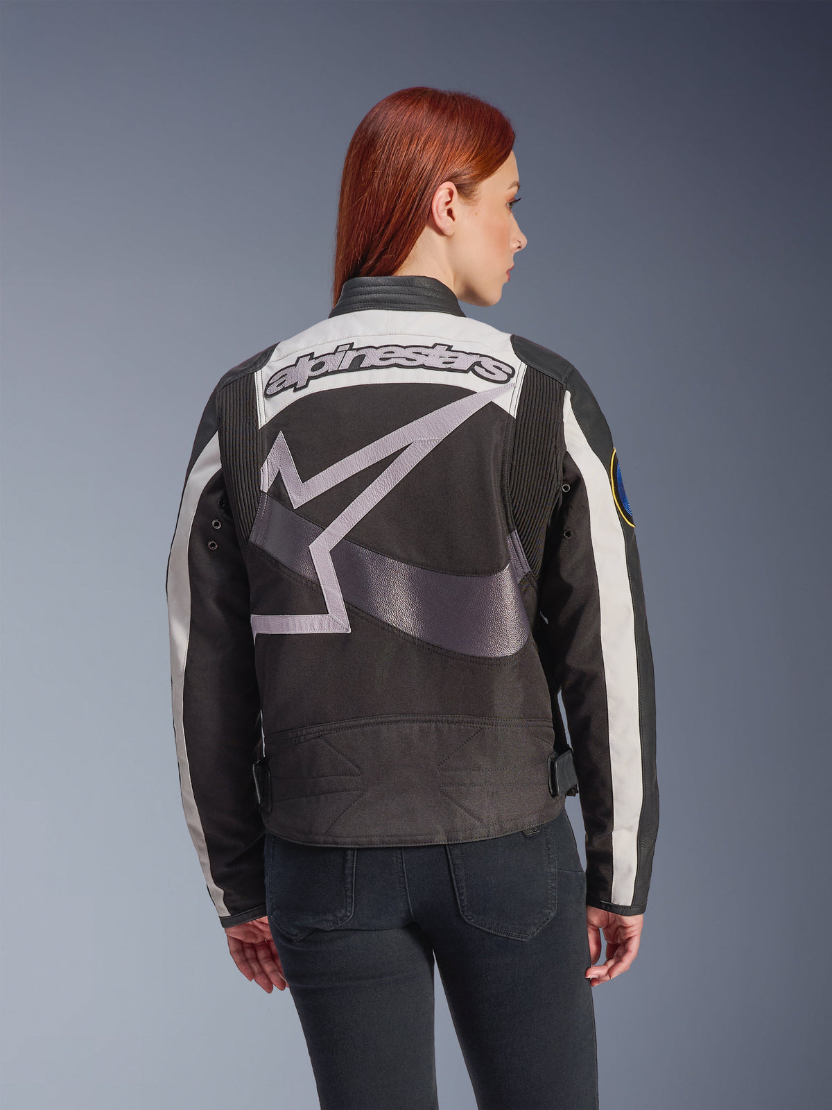 Damska kurtka Alpinestars Idles, czarna i w kolorze złamanej bieli, widok z tyłu z dużym szarym logo gwiazdy i napisem Alpinestars, noszona przez rudowłosą modelkę, miejska kurtka motocyklowa w stylu vintage z panelami akordeonowymi