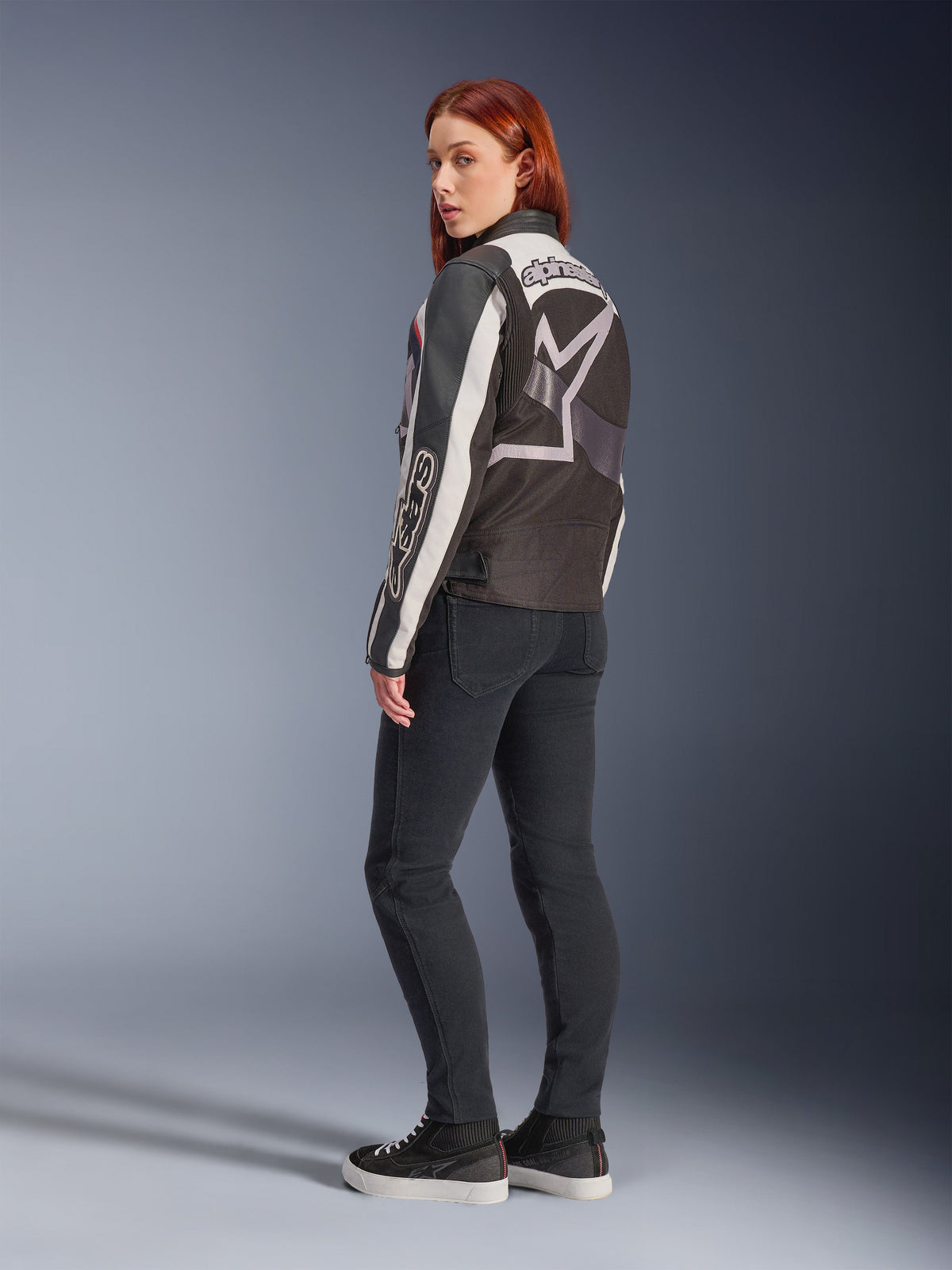 Kobieta w kurtce Alpinestars Idles, widok z tyłu, kolorystyka Off White, Flame Red i czarna, z dużym logo gwiazdy na plecach i napisem Alpinestars na rękawie, zestawiona z czarnymi spodniami typu slim-fit i sneakersami, miejska odzież motocyklowa dla kobiet