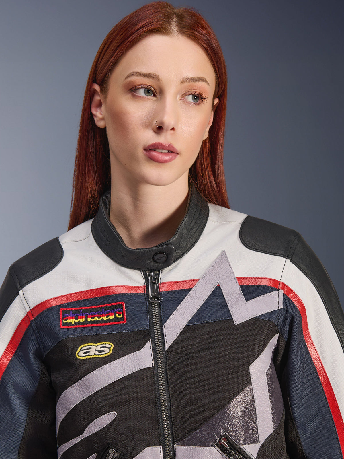 Kurtka damska Alpinestars Idles, zbliżenie na rudowłosą modelkę, kolorystyka Off White, Flame Red i czarna, inspirowany stylem vintage design wyścigowy z naszywkami z logo i szarymi geometrycznymi grafikami gwiazdy, miejska odzież motocyklowa.
