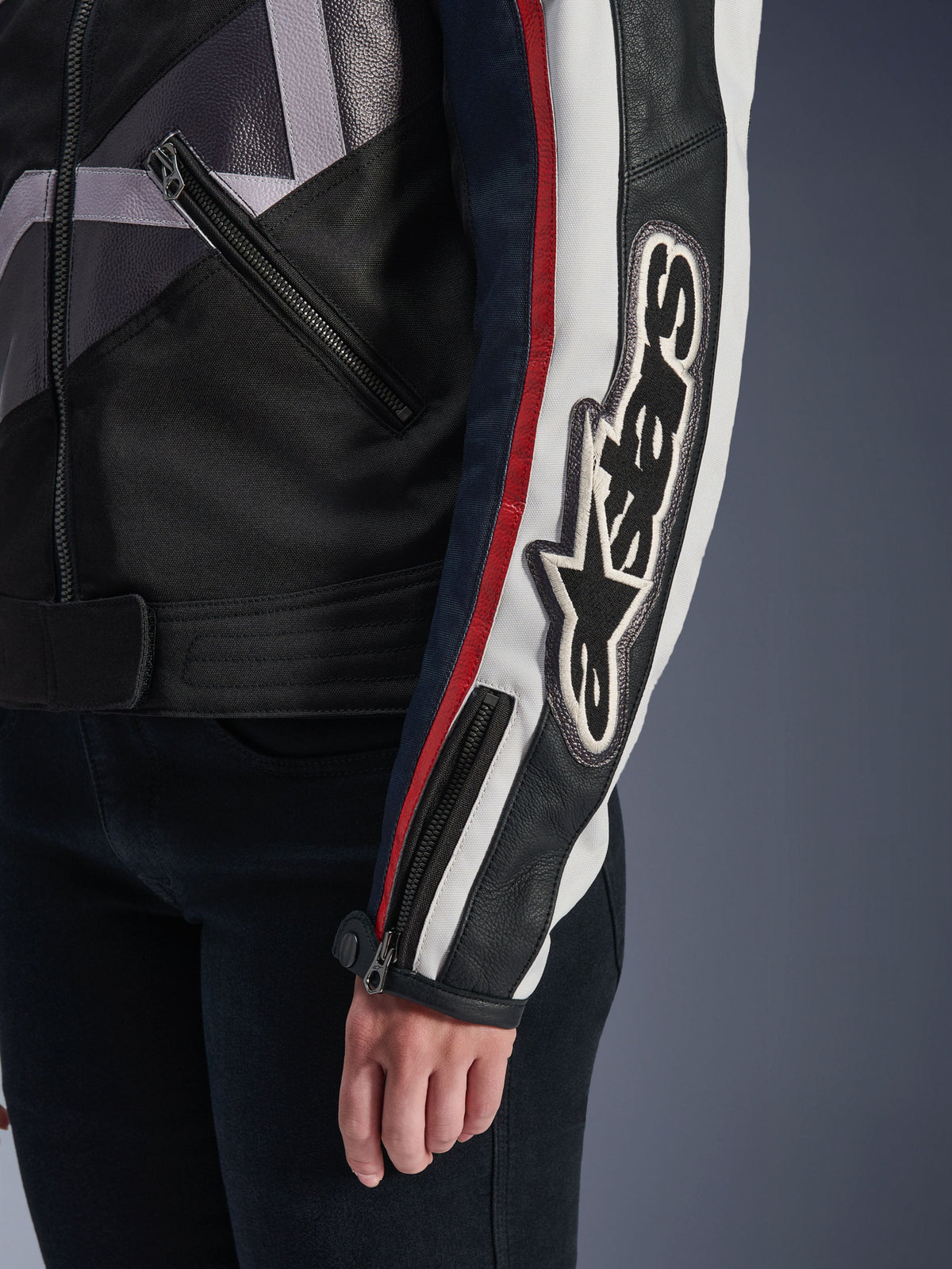 Kurtka damska Alpinestars Idles, miejska kurtka motocyklowa, kolor Off White Flame Red Black, zbliżenie na rękaw z dużym haftowanym logo Alpinestars, czerwone i białe wyścigowe paski, czarna konstrukcja tekstylno-skórzana i metalowe detale zamków.