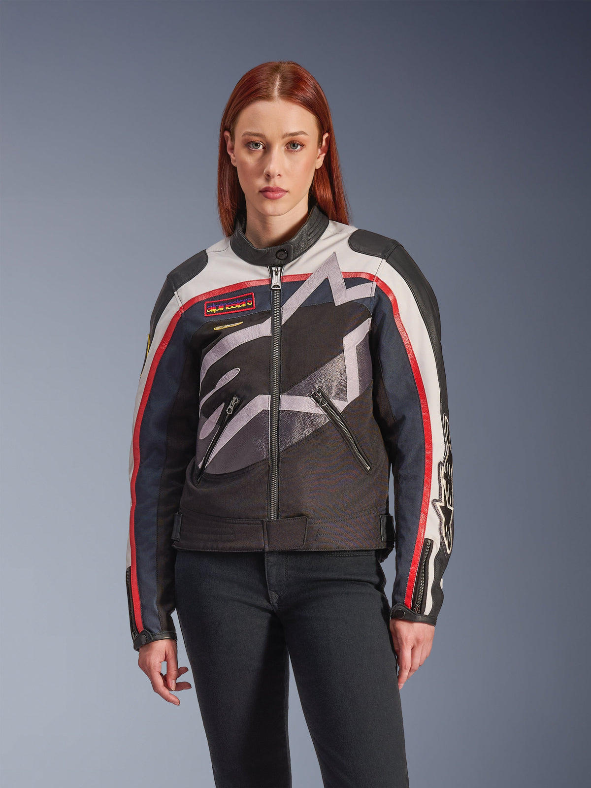 Alpinestars Idles Jacket, damska kurtka motocyklowa, w kolorach złamanej bieli, czerwieni i czerni, z dużym stylizowanym logo gwiazdy, naszywkami na piersi w stylu vintage, wysokim kołnierzem, kieszeniami na zamek i paskami na rękawach, noszona przez modelkę z ciemnymi dżinsami.