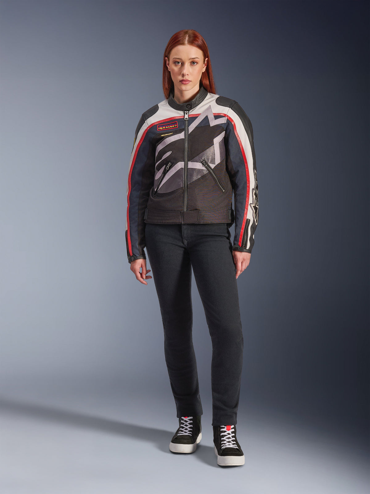 Modelka ubrana w damską kurtkę motocyklową Alpinestars Idles, kolorystyka Off White Flame Red i czarny, z dużym, oplatającym logo gwiazdy i brandingiem inspirowanym stylem vintage, zestawiona z ciemnymi spodniami motocyklowymi slim-fit i czarnymi wysokimi sneakersami, miejska współczesna odzież motocyklowa