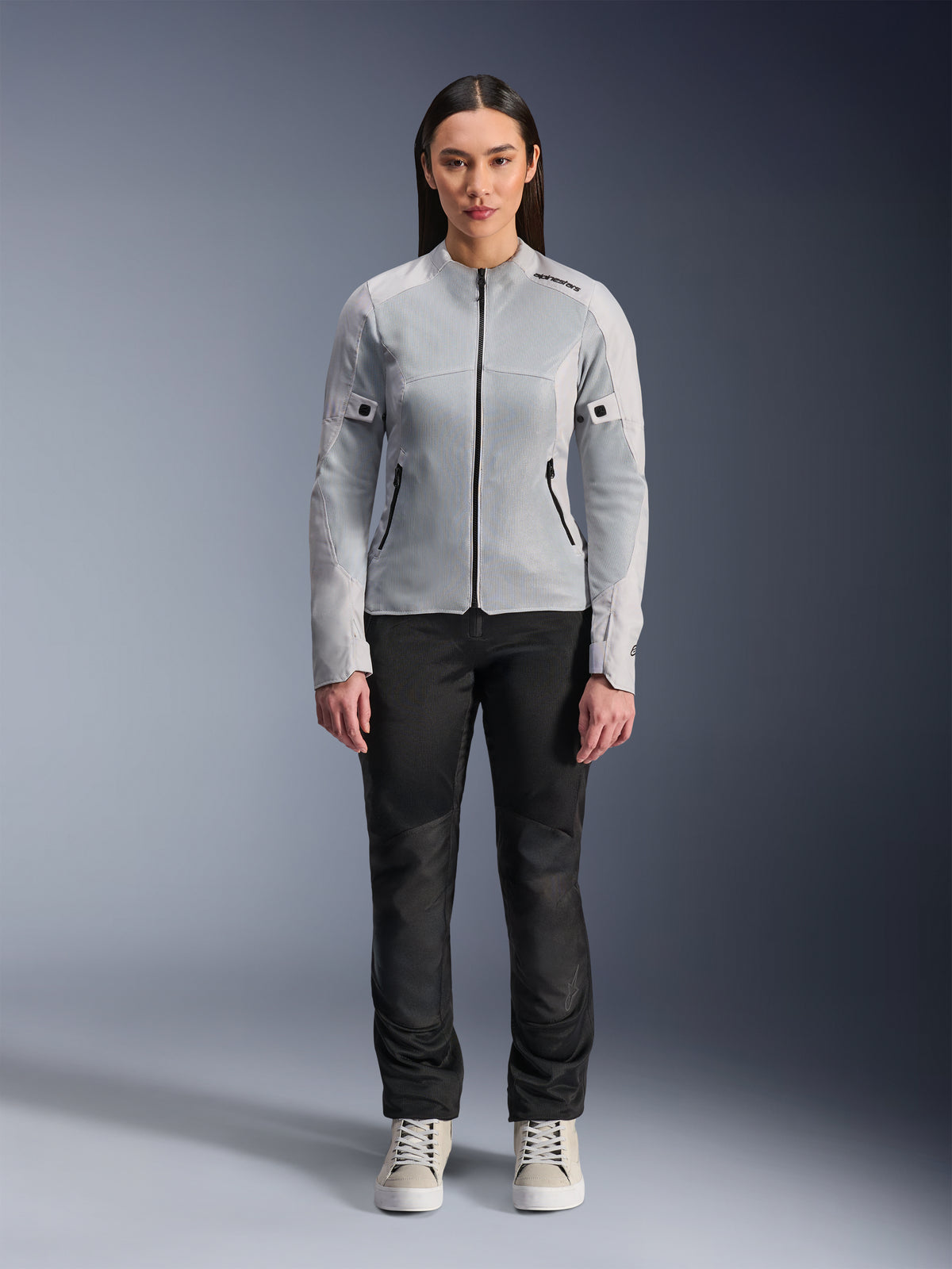 Kobieta w kurtce Alpinestars Stella C-1 Air w kolorze srebrnym, widok z przodu, miejska motocyklowa kurtka tekstylna z panelami z siatki technicznej, zestawiona z czarnymi spodniami i białymi trampkami, nowoczesna odzież motocyklowa